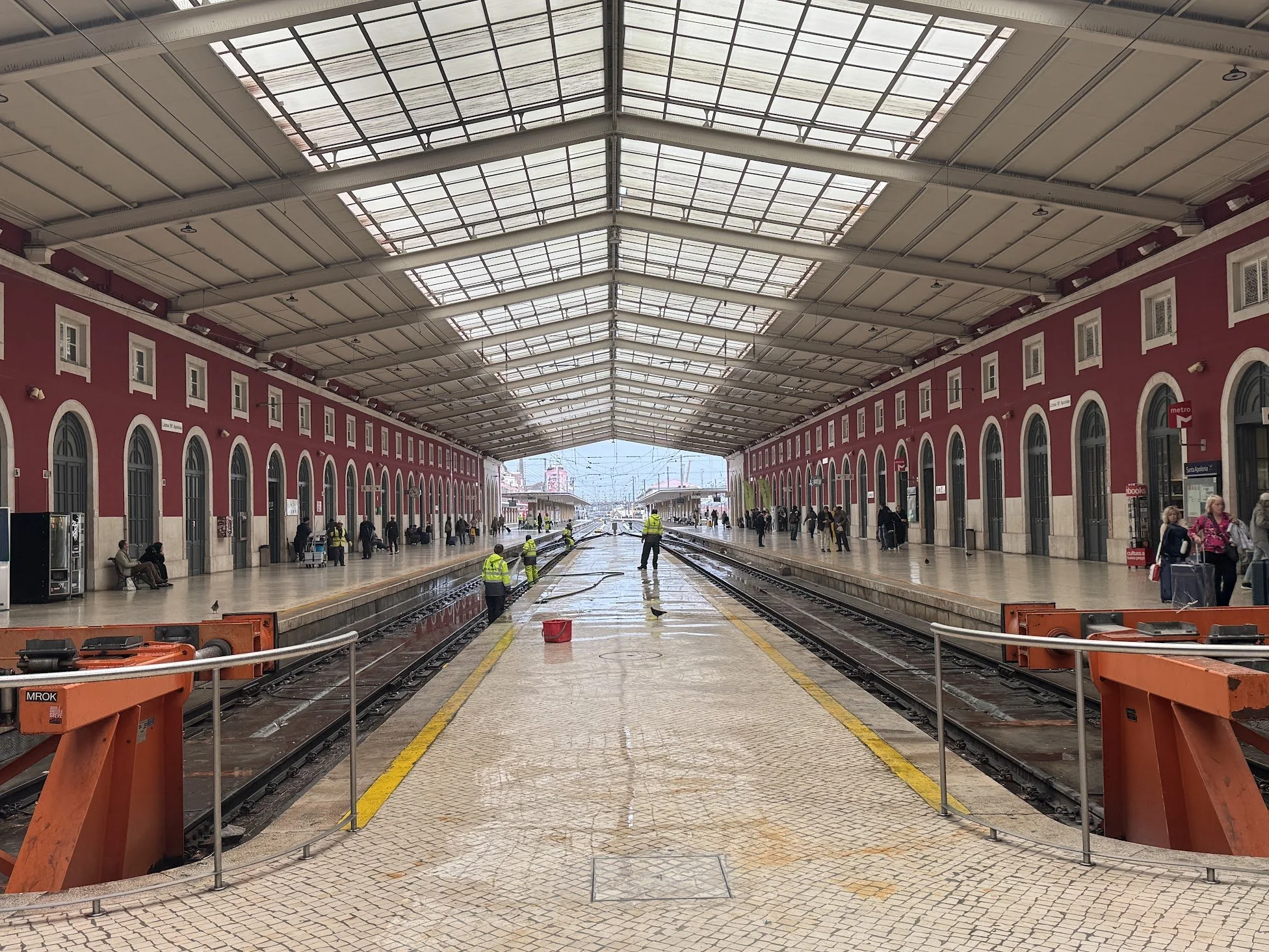 View of a train station platform during cleaning, with construction barriers, workers in safety vests, and an arched roof overhead.