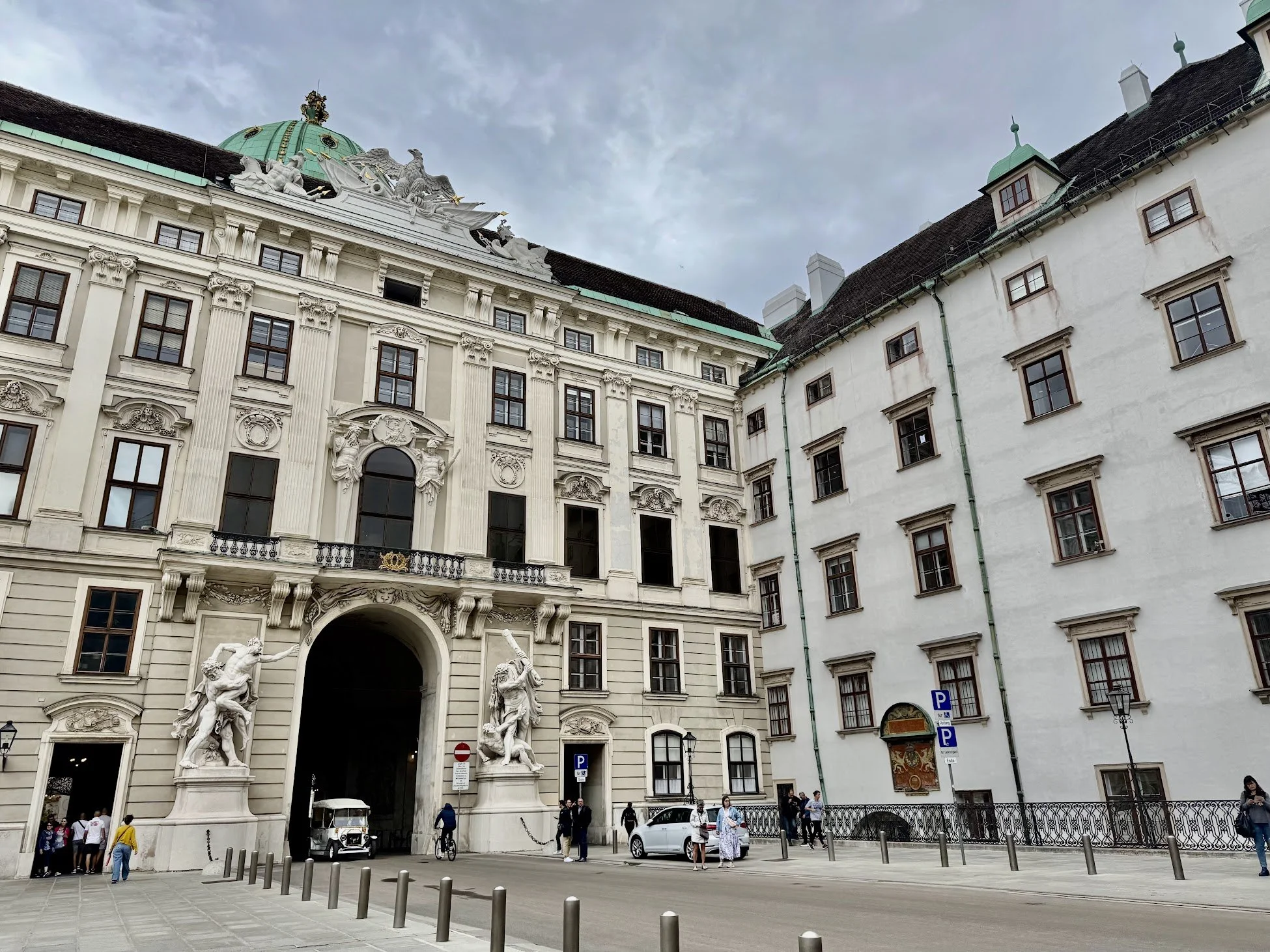 A historic building with ornate architectural details and sculptures at the entrance, situated in a busy urban area with people and cars in front.