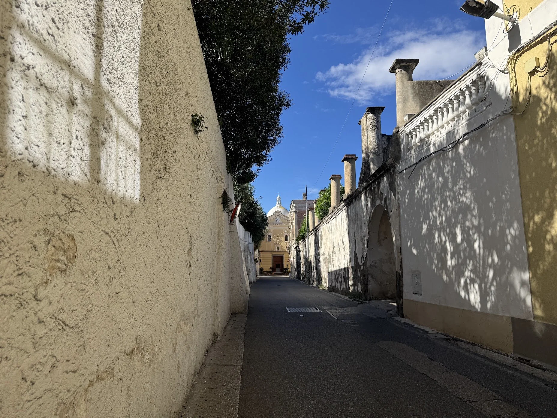 A narrow street flanked by tall, aged white and yellow walls with textured surfaces. Shadows from trees cast patterns on the right wall. In the distance, there is a church with a domed roof and a decorative façade, under a bright blue sky with some c