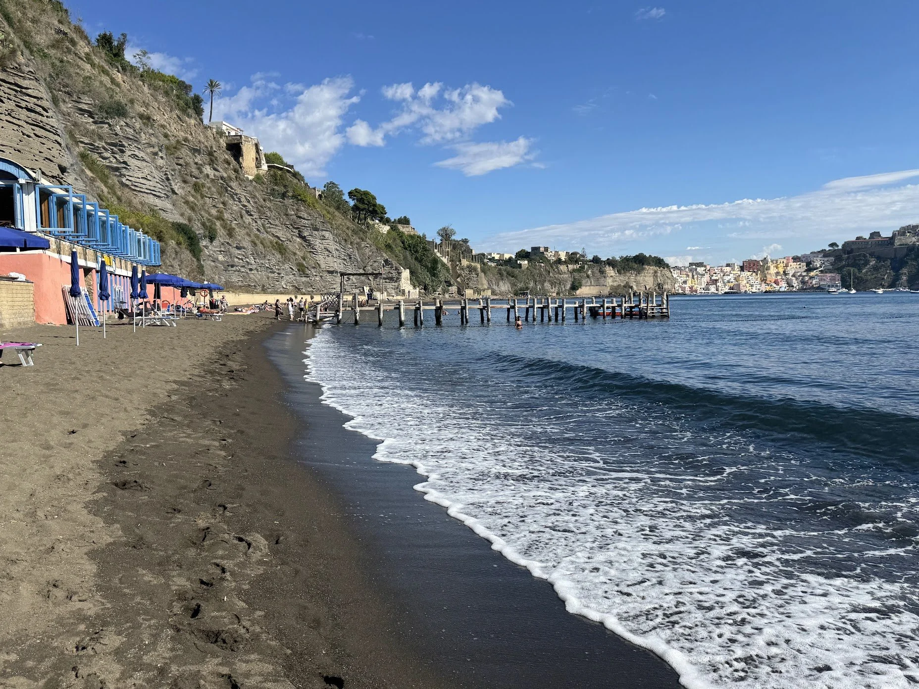 Beach with dark sand, blue umbrellas, and a wooden pier extending into the water, with a hillside and colorful buildings in the background under a partly cloudy sky.