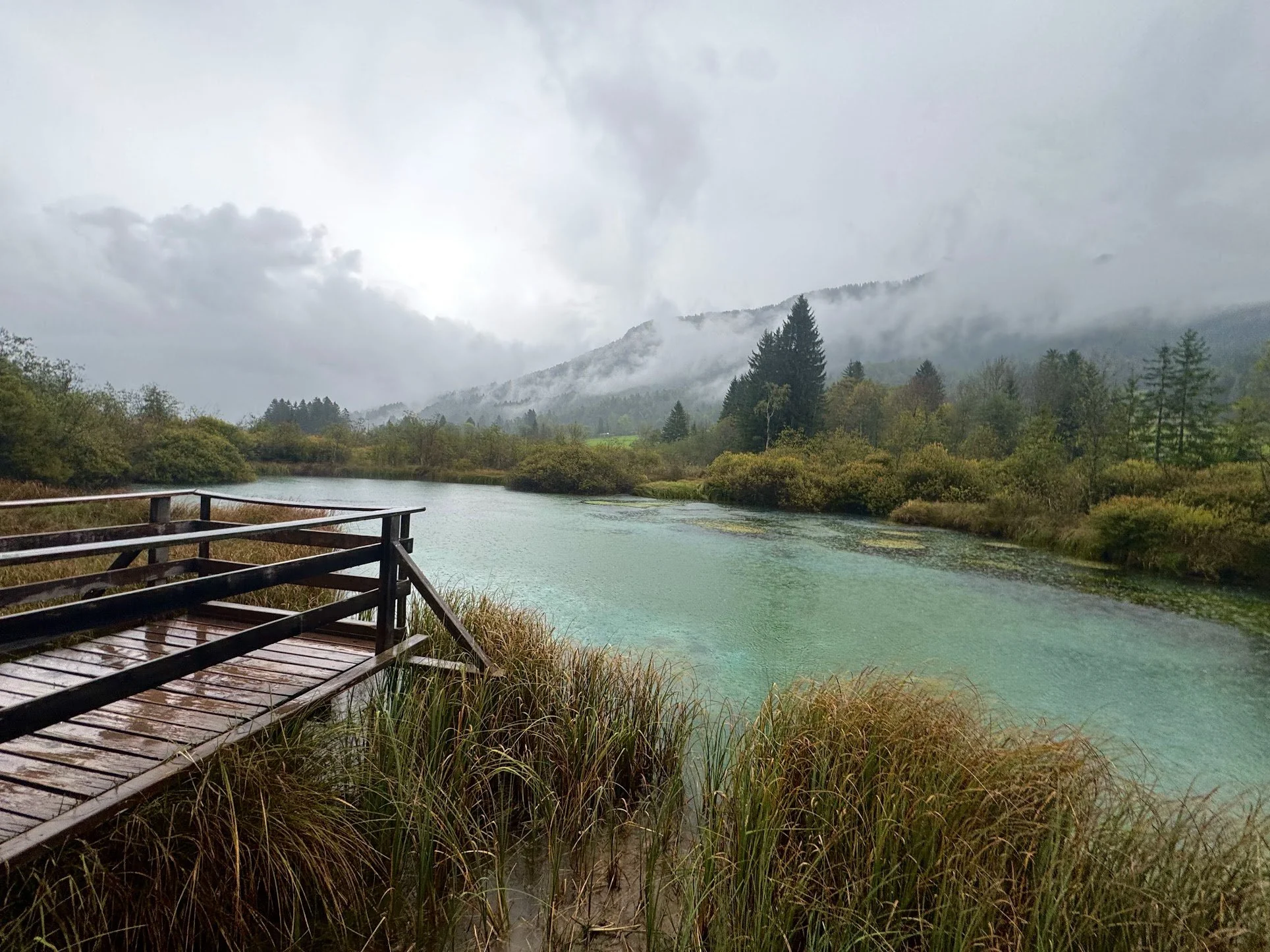 A wooden deck overlooking a calm river surrounded by lush green foliage. In the background, forested hills are shrouded in mist and clouds, creating a serene, misty mountain landscape.