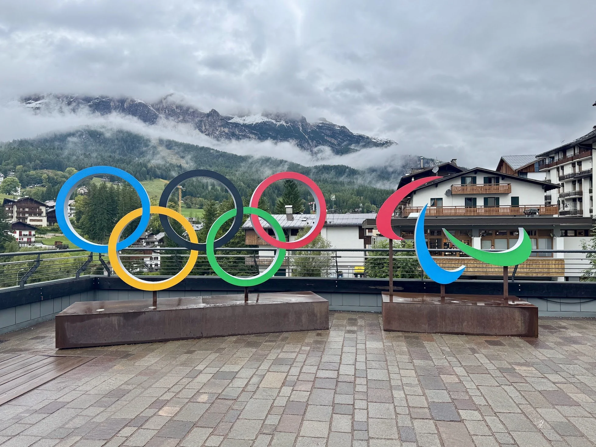 Olympic rings sculpture on a rooftop with mountains and houses in the background, and cloudy skies overhead.