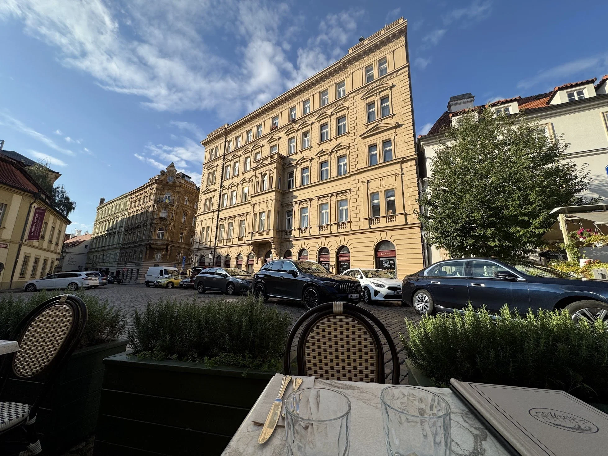 View from an outdoor cafe seating area looking at historic buildings across a cobblestone street with parked cars, trees, and a partly cloudy sky.
