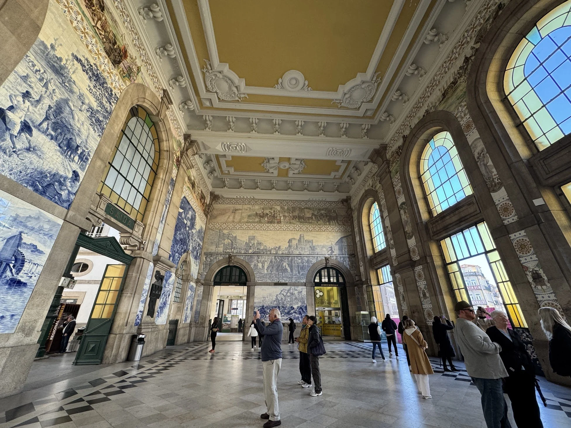 Interior of a historic train station with ornate white and yellow ceiling, arched stained glass windows, decorative wall murals, and people waiting or taking photos.