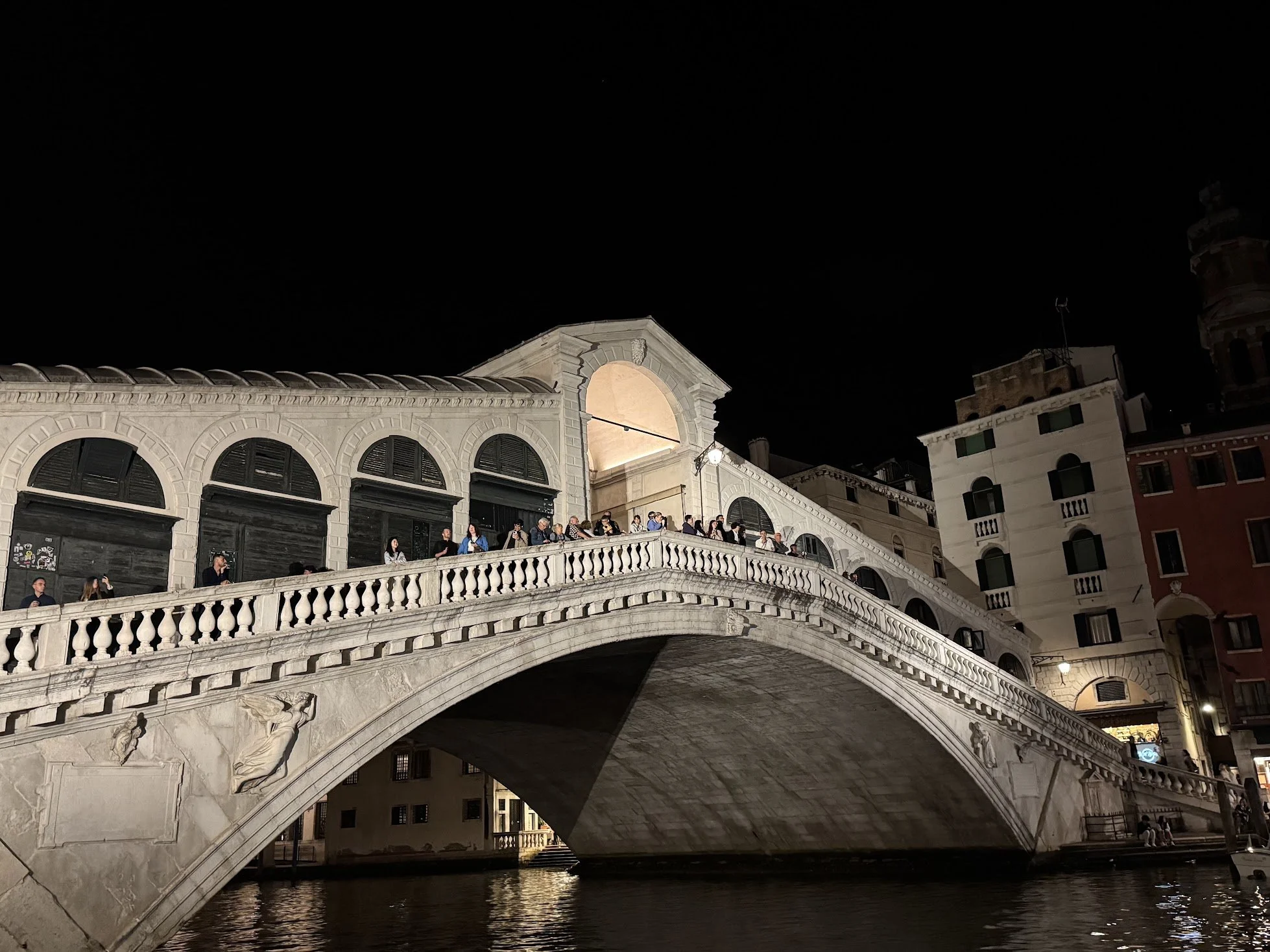 Night view of a historic arched stone bridge with a staircase leading up to a plaza with people gathered, and buildings in the background.
