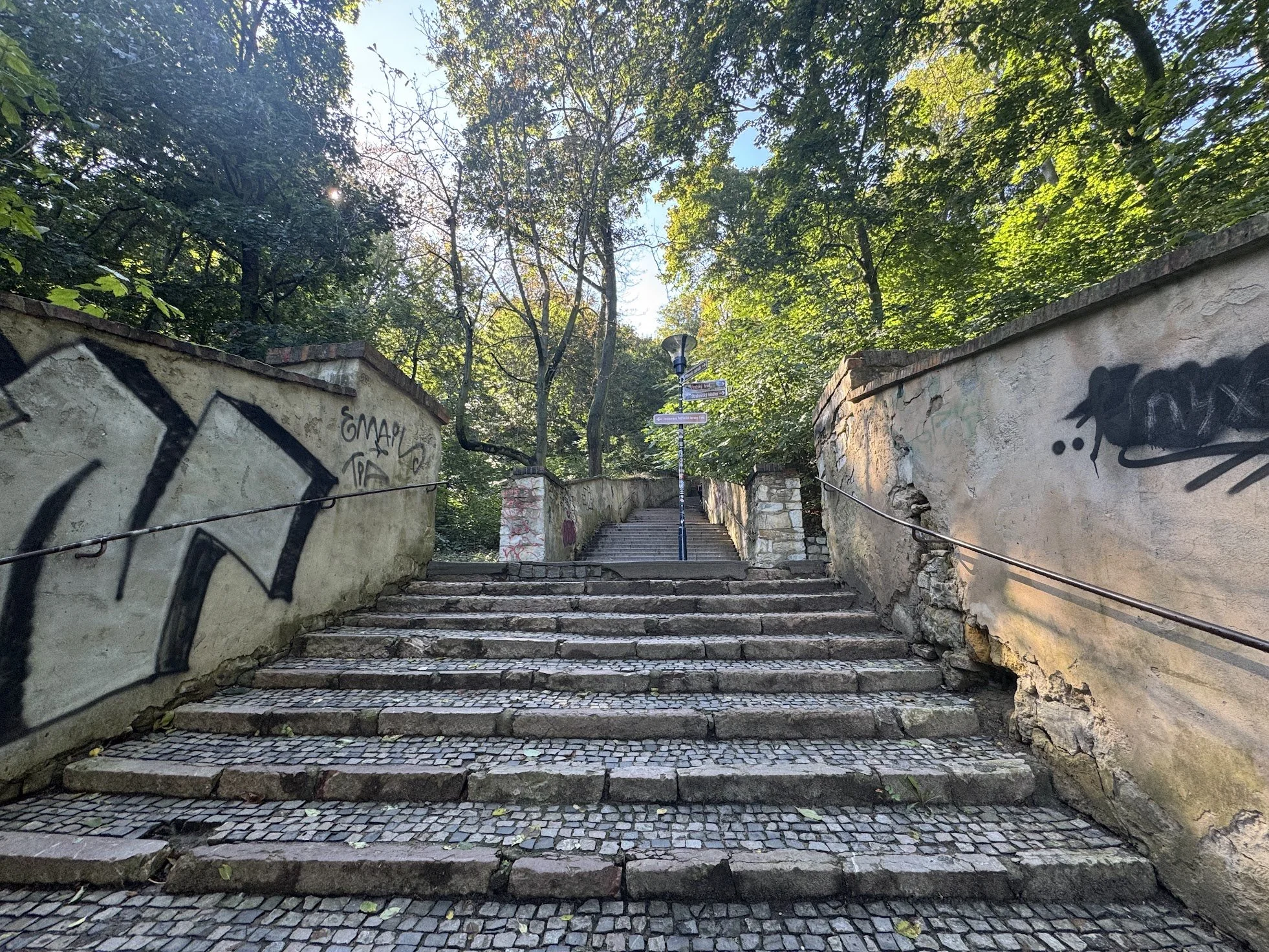 Stone outdoor staircase with graffiti on the walls on either side, surrounded by trees with green leaves and a street sign post with multiple signs, under a bright sky.