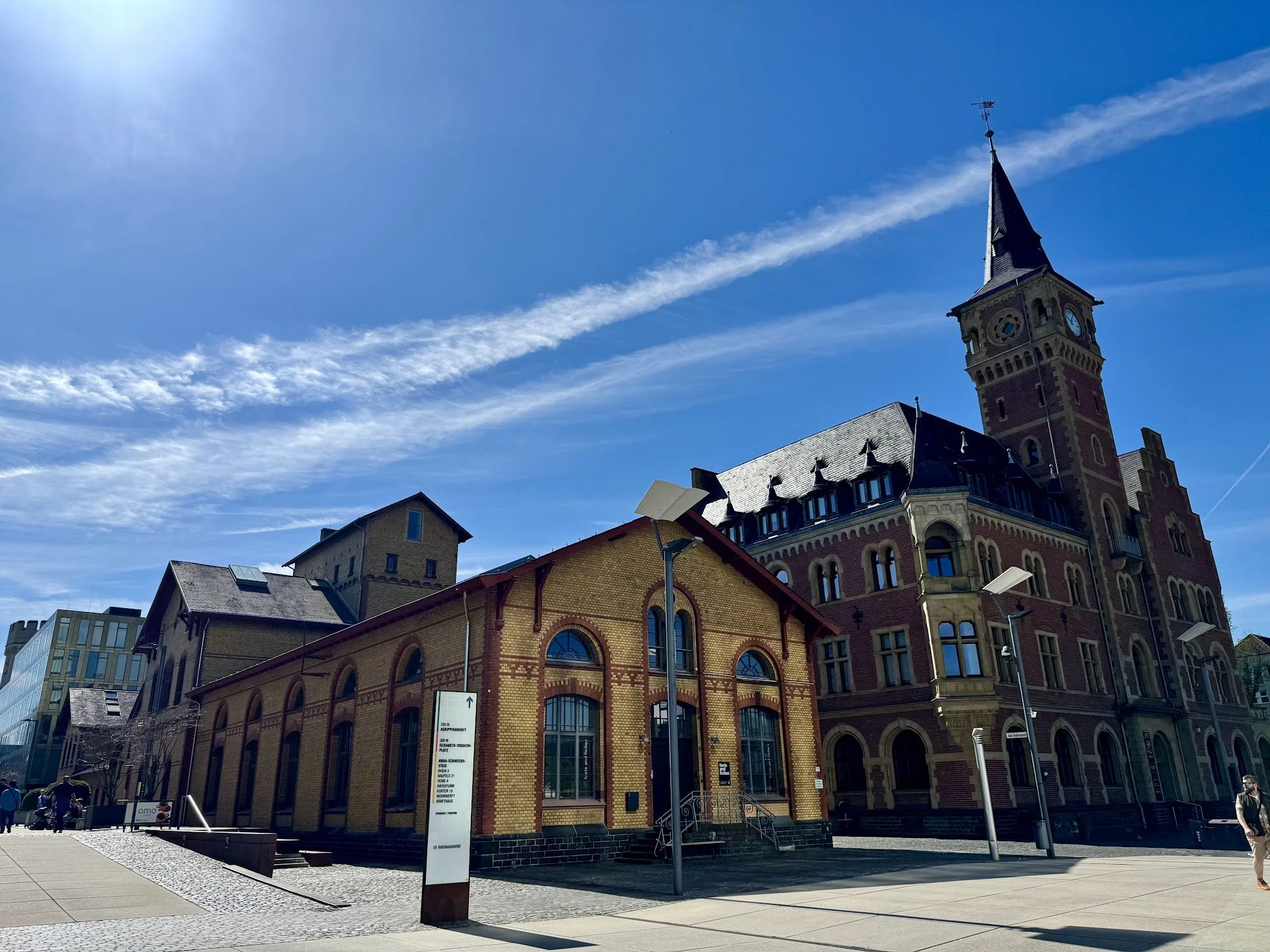 A historic brick building with a clock tower and a pointed steeple, set against a bright blue sky with streaks of clouds. People are walking nearby, and modern streetlights and signage are in the foreground.