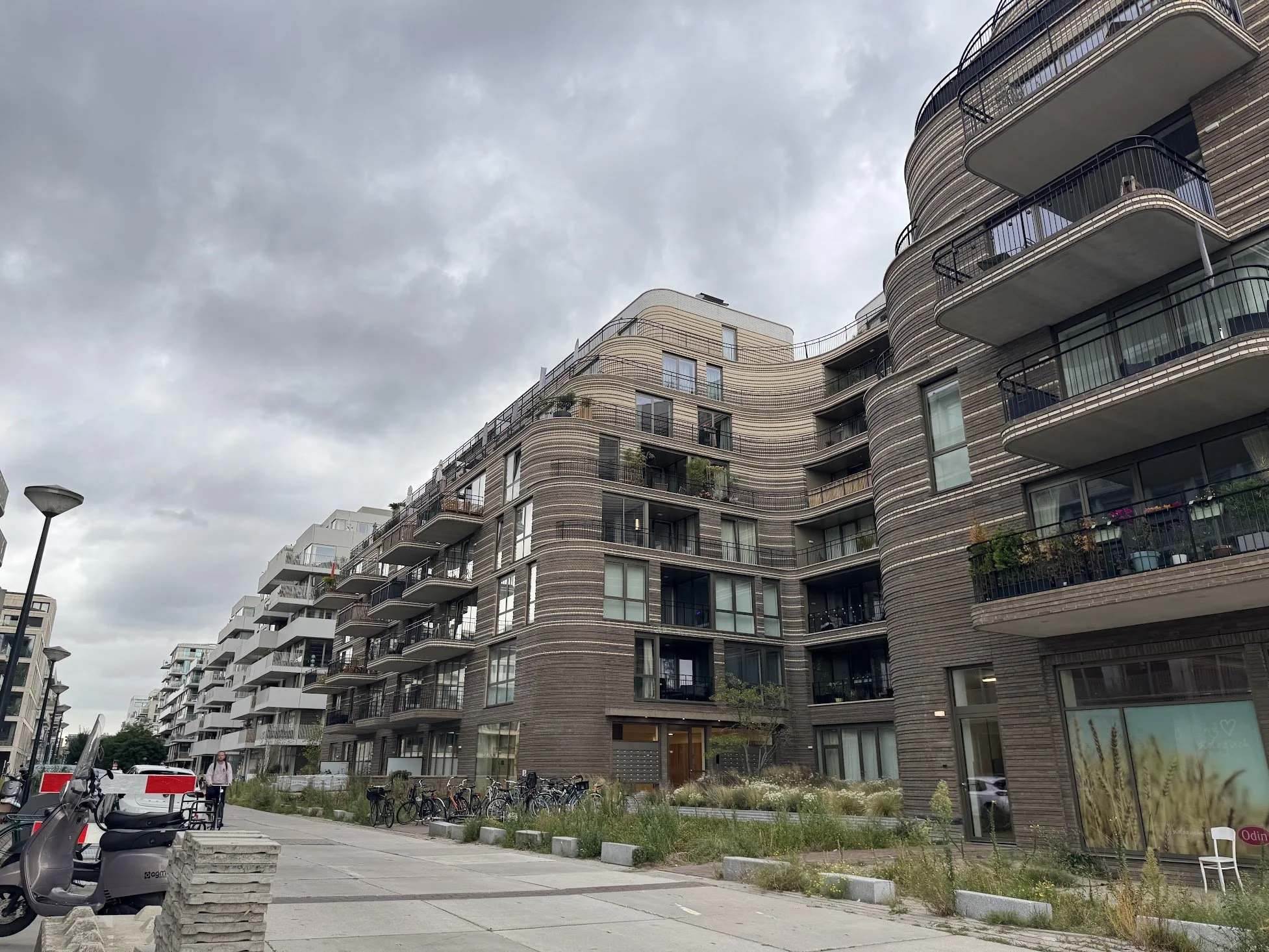 Modern multi-story apartment building with rounded balconies and a brick exterior, overcast sky in an urban setting with bicycles and a sidewalk in the foreground.