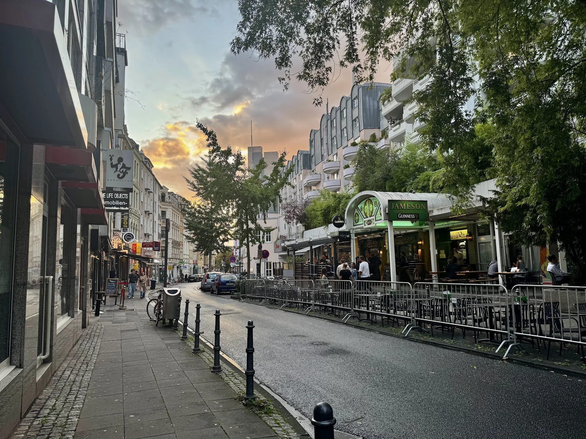 City street scene at sunset with buildings, trees, sidewalk, parked cars, and outdoor seating area with people.