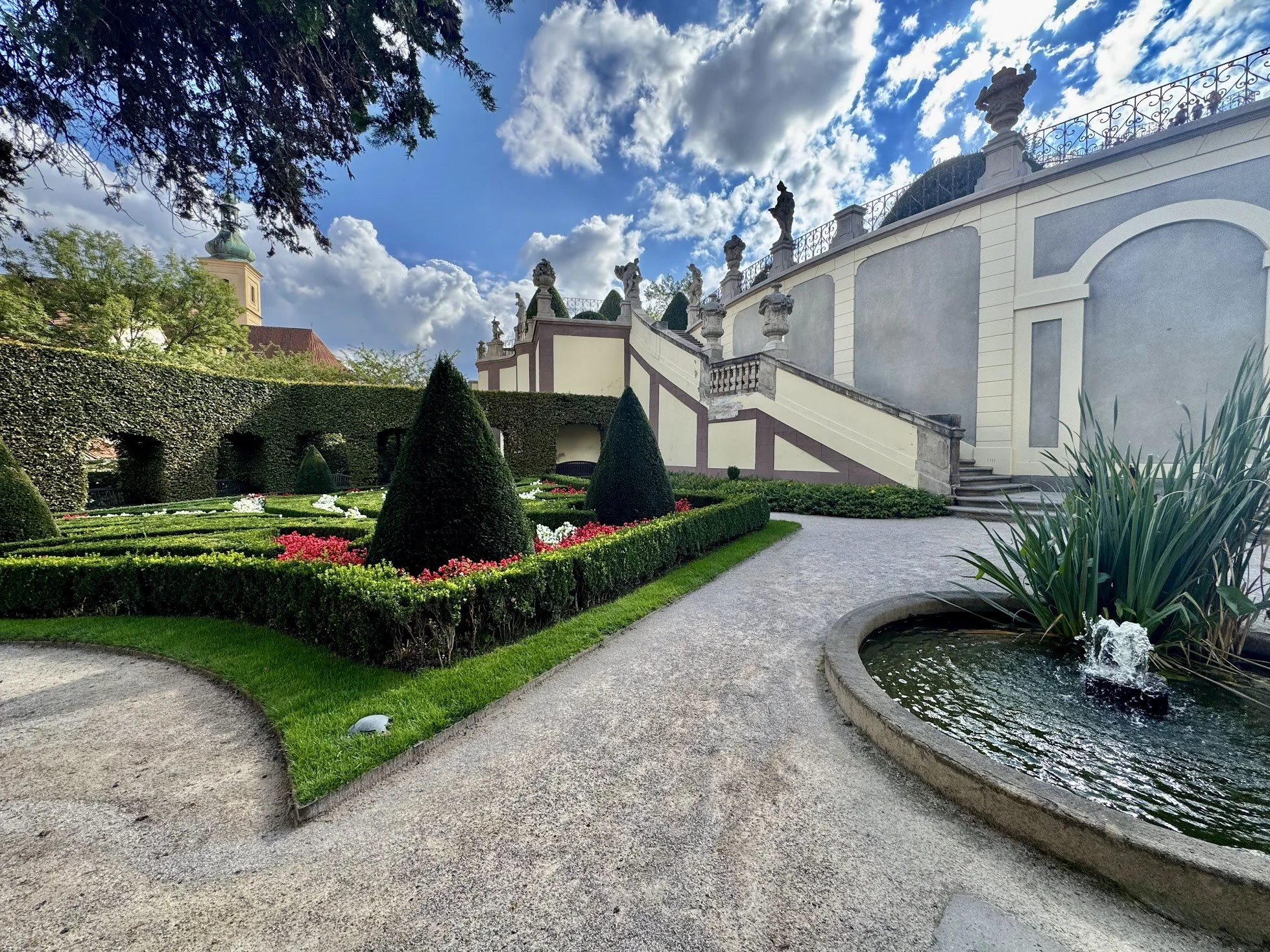 A landscaped garden with neatly trimmed hedges, colorful flower beds, and a small pond with fountain, adjacent to a large, ornate building with white walls and decorative statues on a staircase, under a partly cloudy sky.