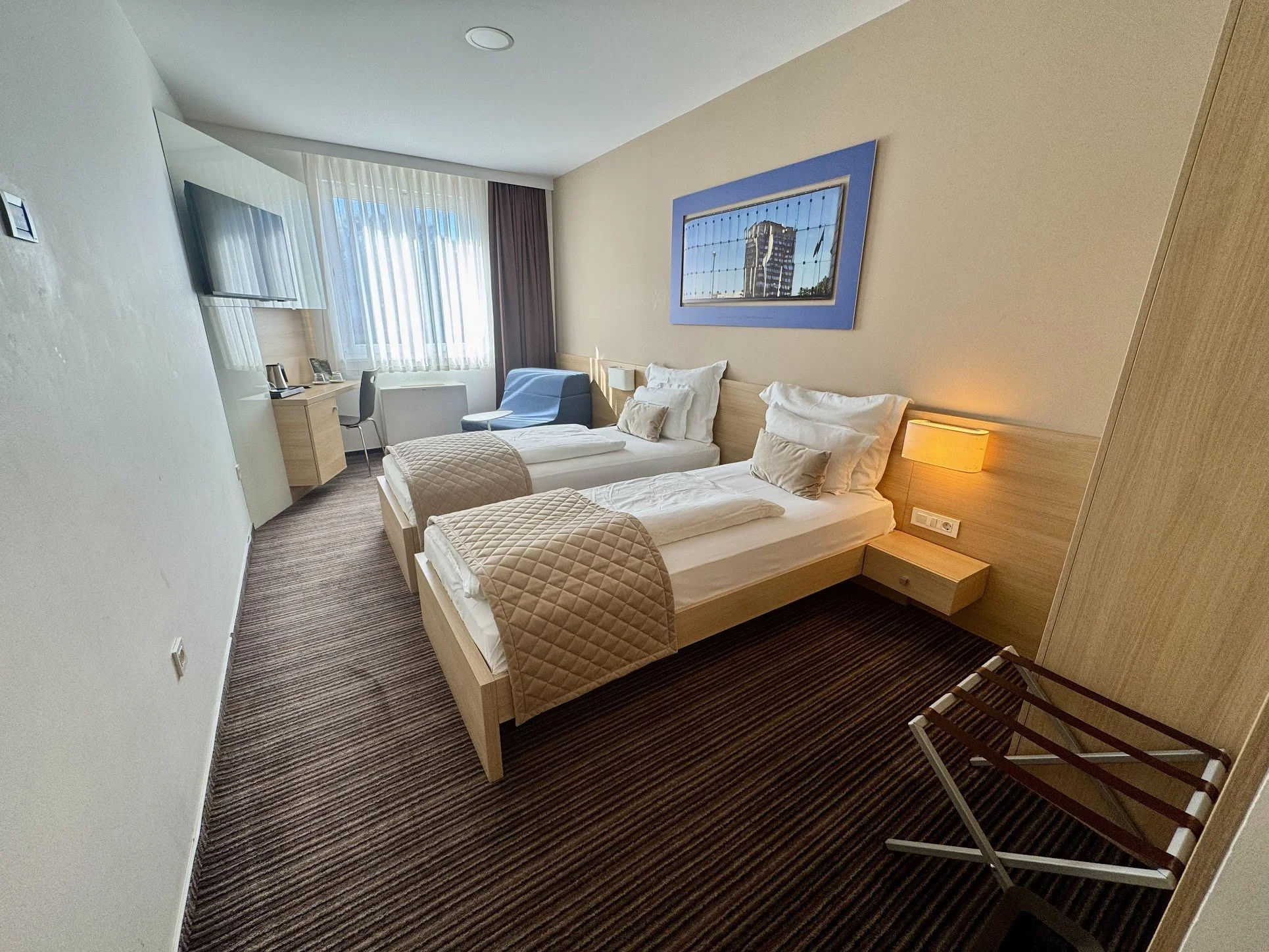 Hotel room with two twin beds, beige walls, window with curtains, framed cityscape picture, desk with telephone and kettle, armchair, wall-mounted TV, and luggage rack.