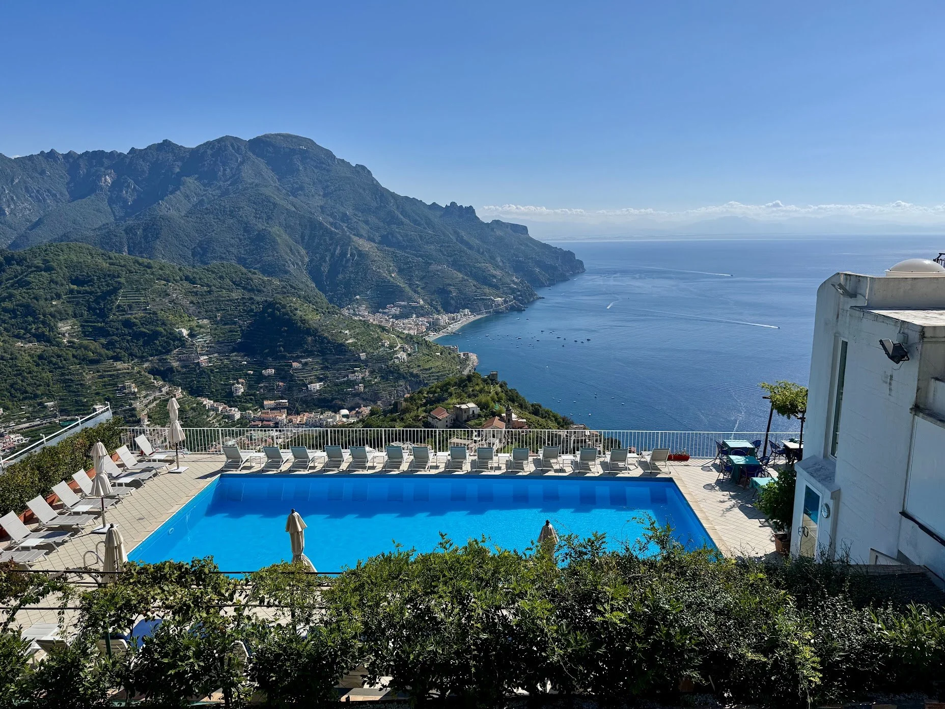A rooftop pool area overlooking a scenic coastline with mountains and blue ocean water, with lounge chairs and umbrellas around the pool.