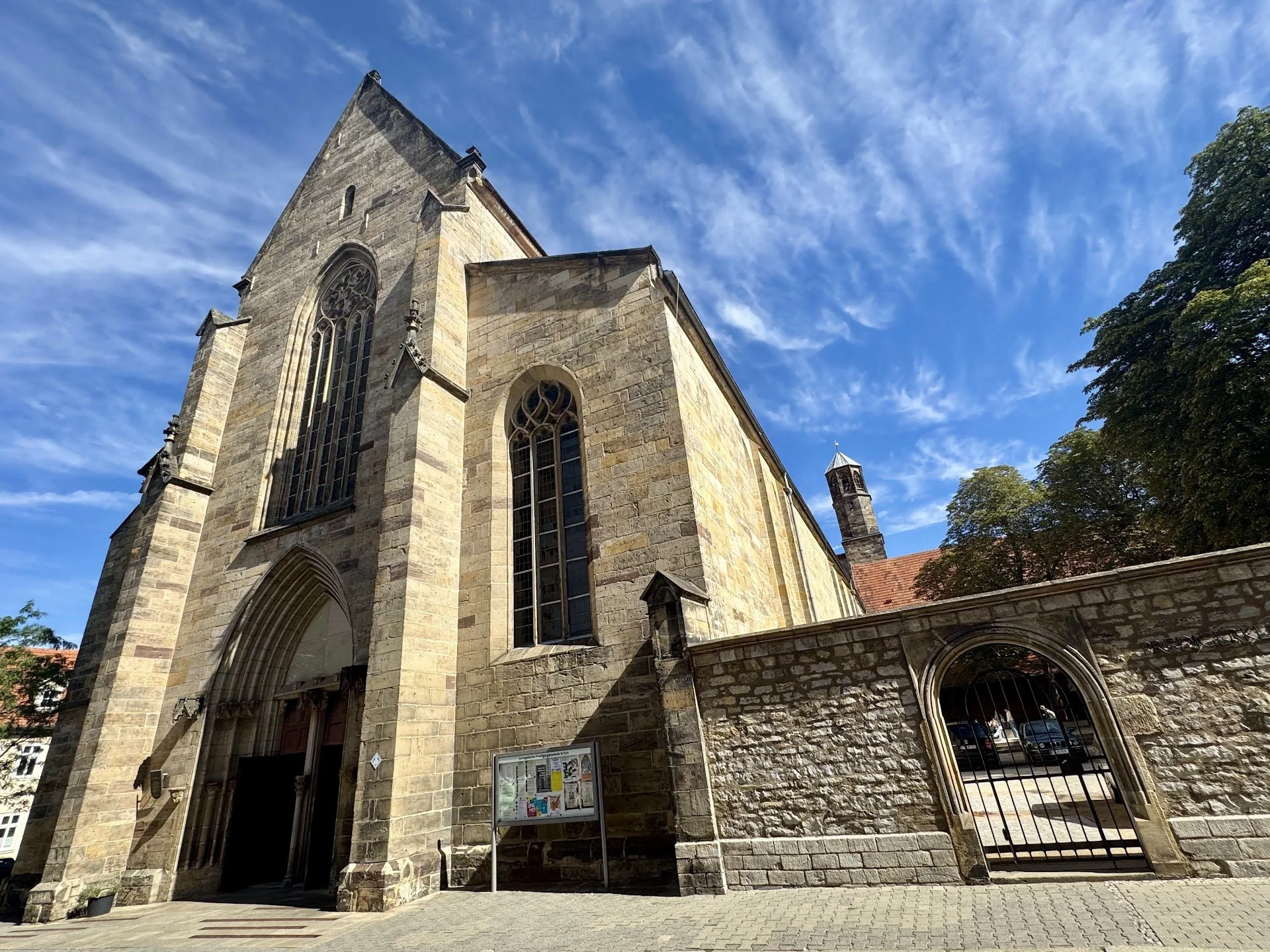 A stone church with large stained glass windows, pointed arch doors, and a small tower with a cross on top, set against a bright blue sky with wispy clouds and green trees.