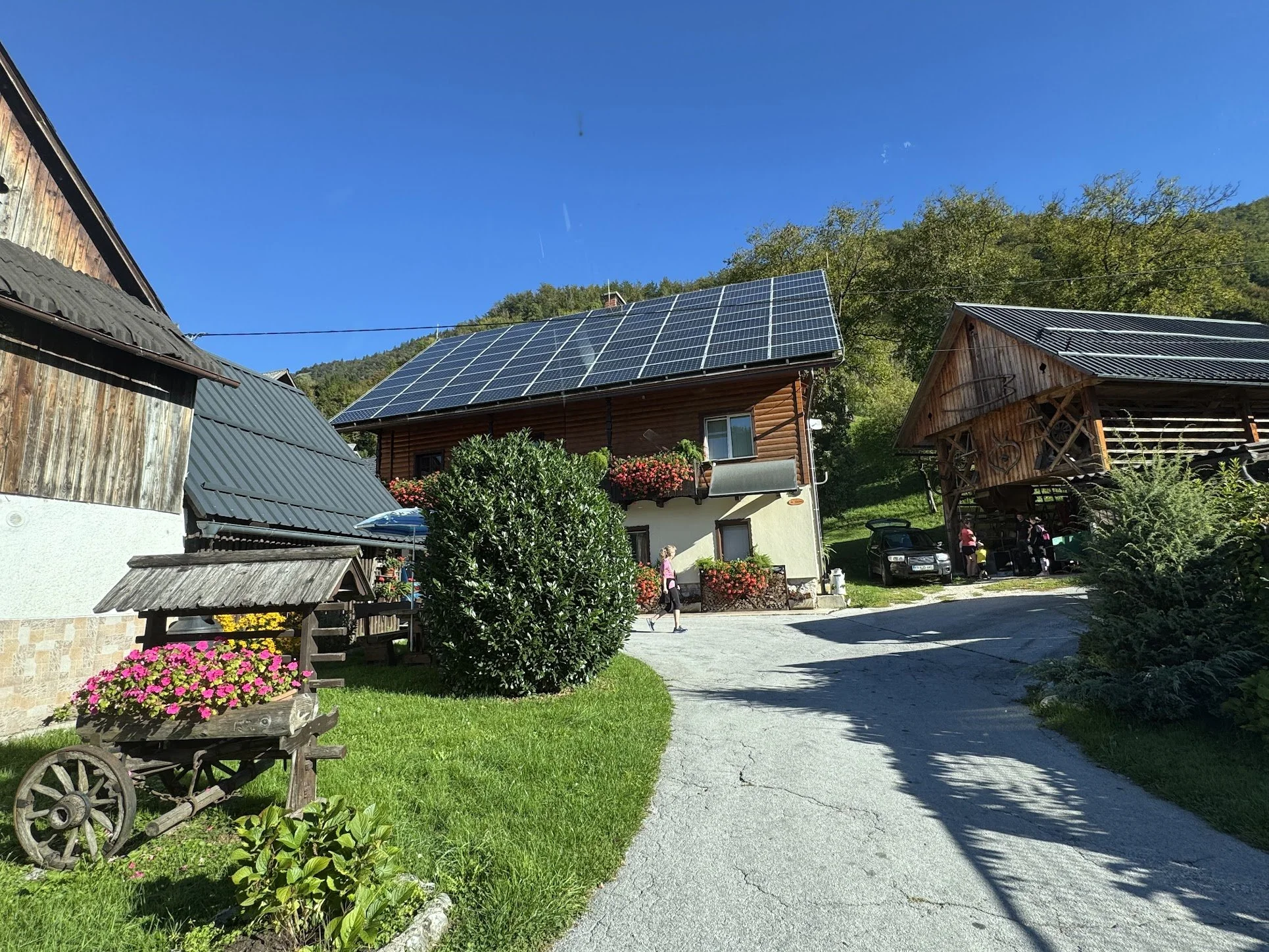A rural scene with a clear blue sky, a house with solar panels on the roof, wooden buildings, colorful flowers, and trees in the background. People are walking on a paved path near parked cars.