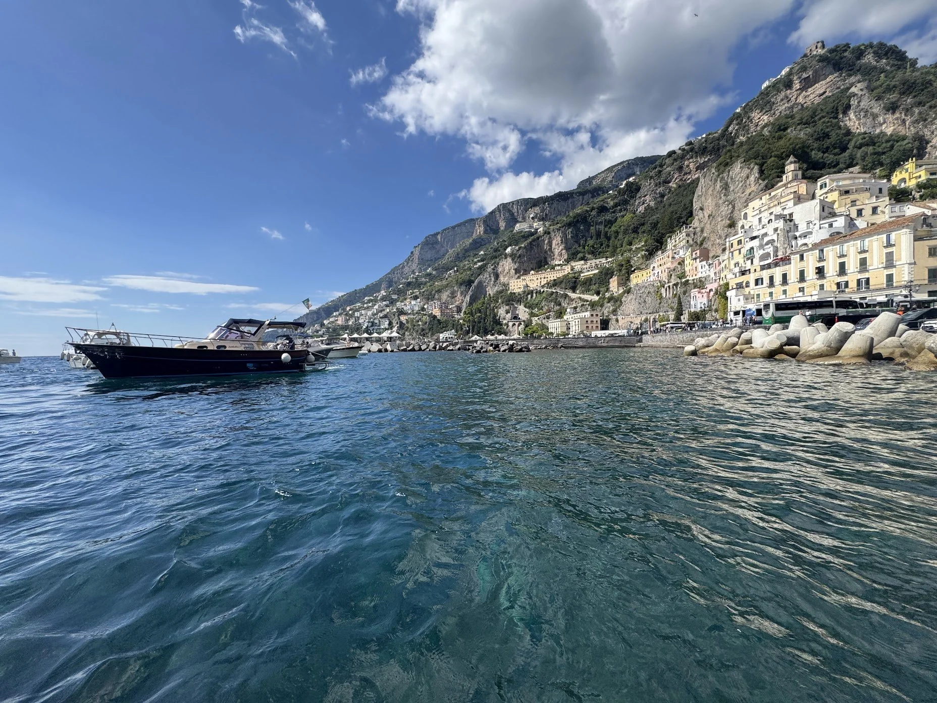 Coastal town with colorful buildings on a hillside next to the sea, boats in the water, rocky shoreline, under a partly cloudy sky.