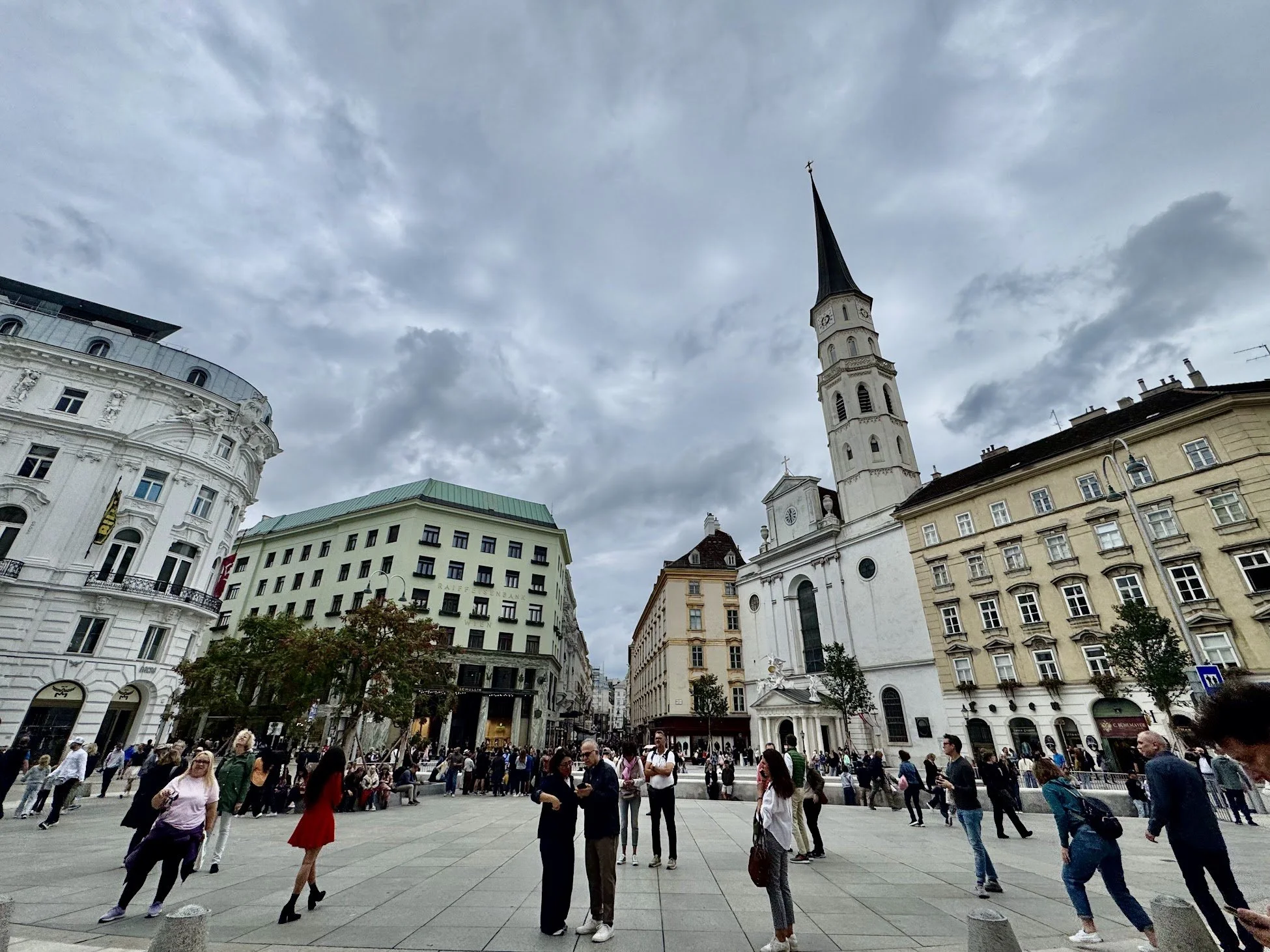 A busy city square with a crowd of people walking and standing. Surrounding buildings include a white ornate building on the left, a green-roofed building in the center, and a church with a tall steeple on the right. The sky is cloudy.