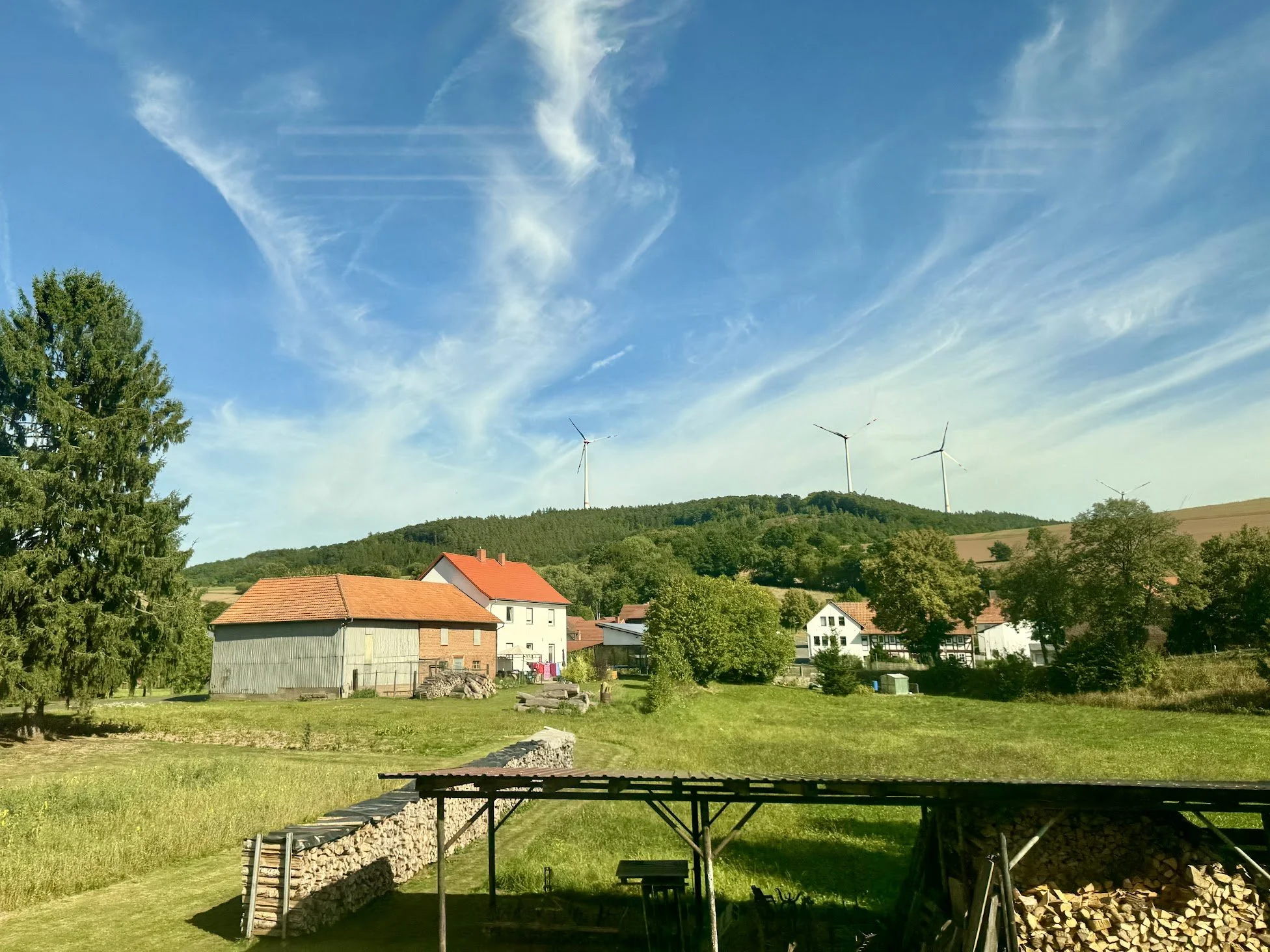 A rural landscape with a house with a red roof, green trees, and a backdrop of wind turbines on a hill under a blue sky with some clouds.
