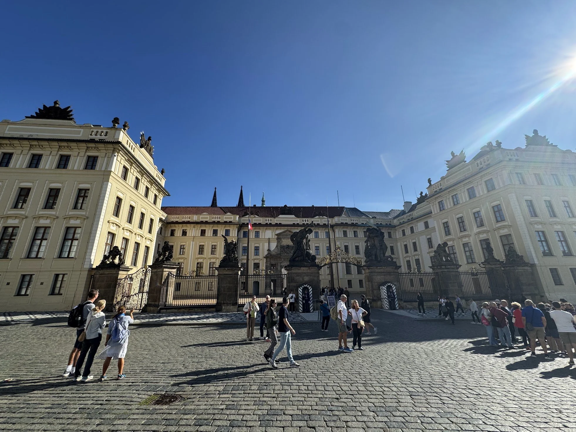 The image shows a large, historic building with statues on its roof and sculptures in front of its entrance. Many people are walking across a cobblestone courtyard in front of the building, with clear blue sky overhead and the sun shining brightly in