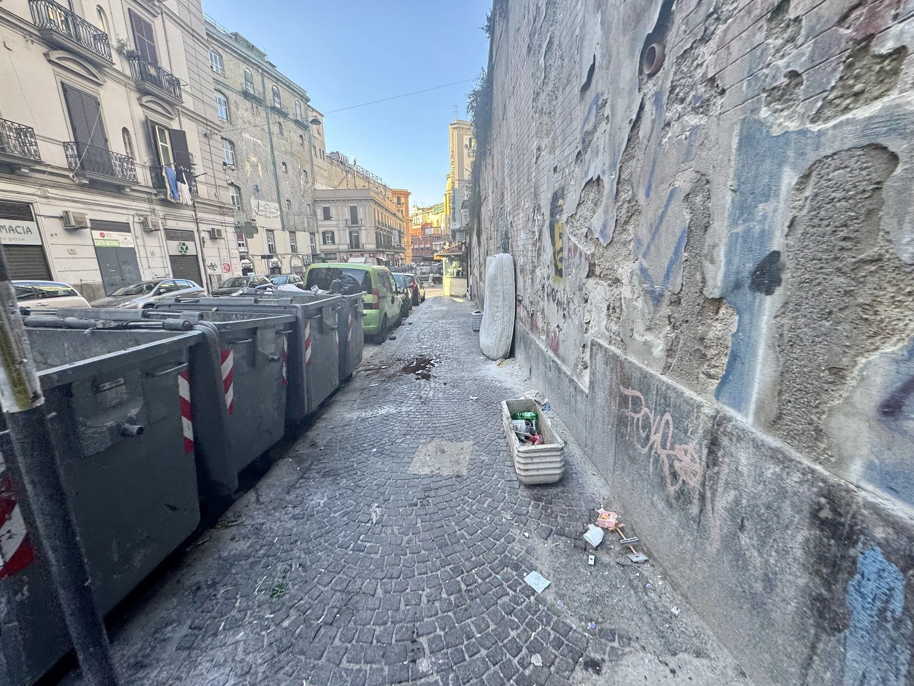 Street scene with parked cars along a narrow sidewalk, graffiti-covered walls, and dumpsters, in an urban area with buildings in the background during daytime.