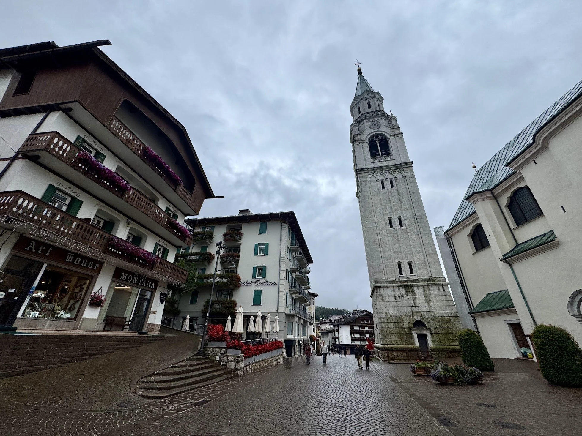 A cobblestone street in a town center with a tall church tower and European-style buildings, some with balconies decorated with flowers, under a cloudy sky.