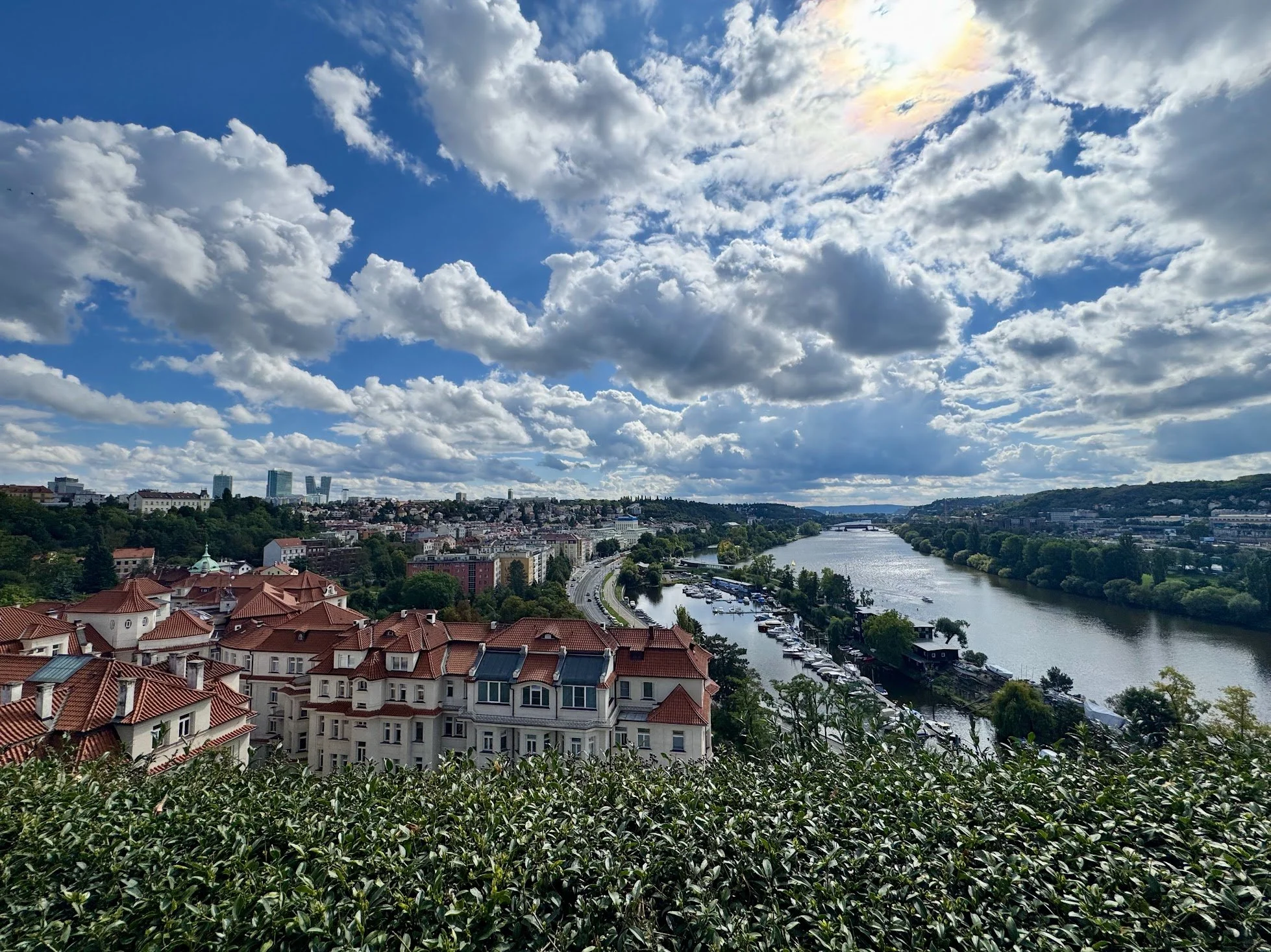Cityscape with red-roofed buildings along a river, under a partly cloudy sky with rainbow and sunlight.