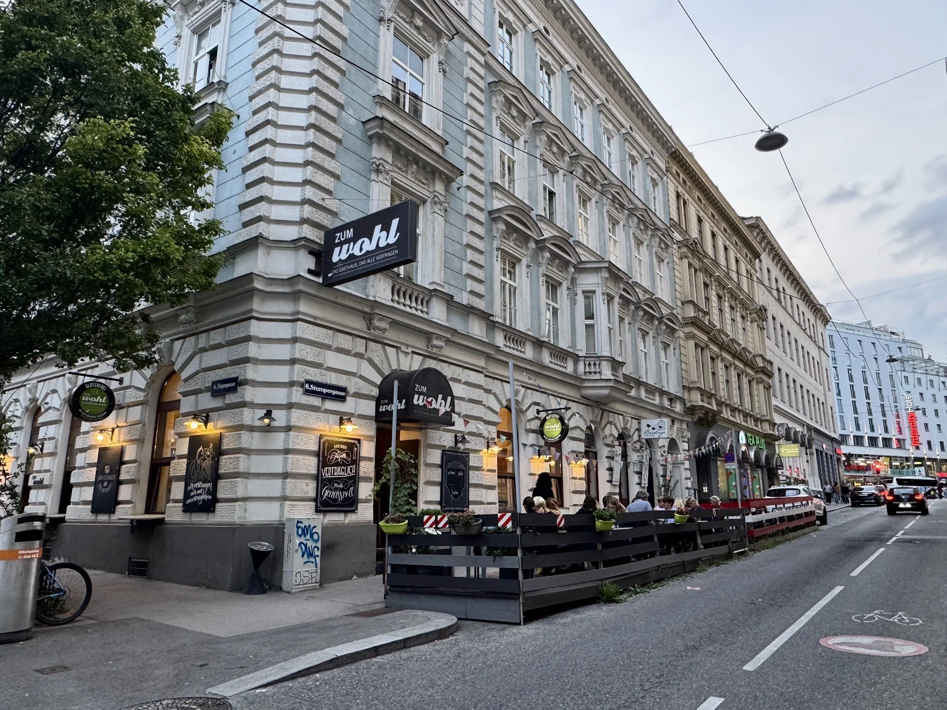 A street corner with a historic light blue and white building housing a bar named 'Zum Wahl.' The building has multiple windows and decorative architectural details. People are seated outside at tables, and there are signs and street signs visible, i