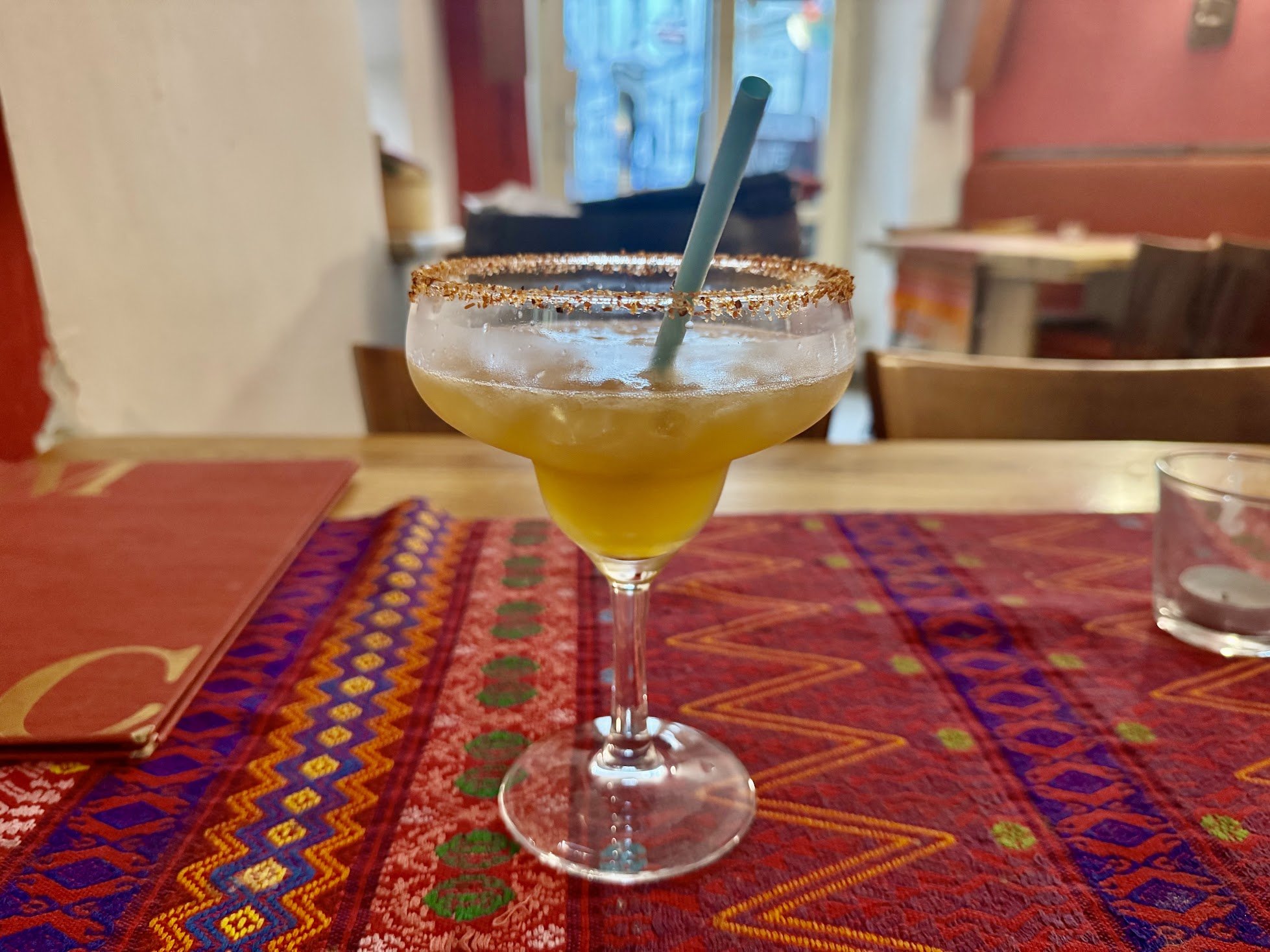 A margarita cocktail in a glass with a salted rim and a green straw on a colorful, patterned table in a restaurant setting.