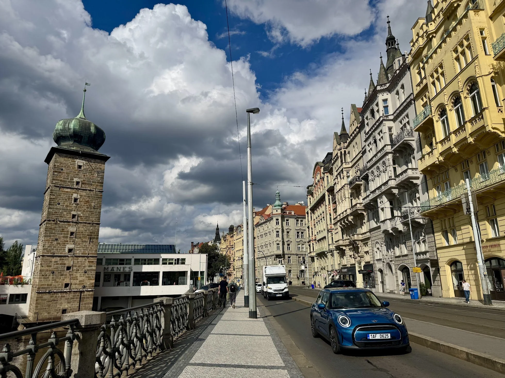 Street view with historic European-style buildings, a tall stone tower with a green, onion-shaped roof, a lamp post, cars parked along the street, and pedestrians walking on the sidewalk.