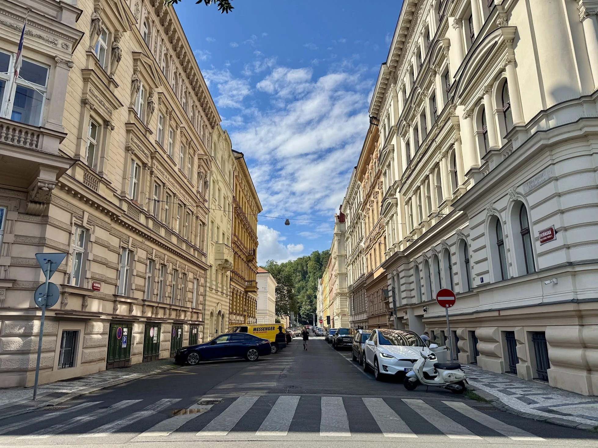 Street view between historic buildings with decorative facades, parked cars, and a clear blue sky with scattered clouds.