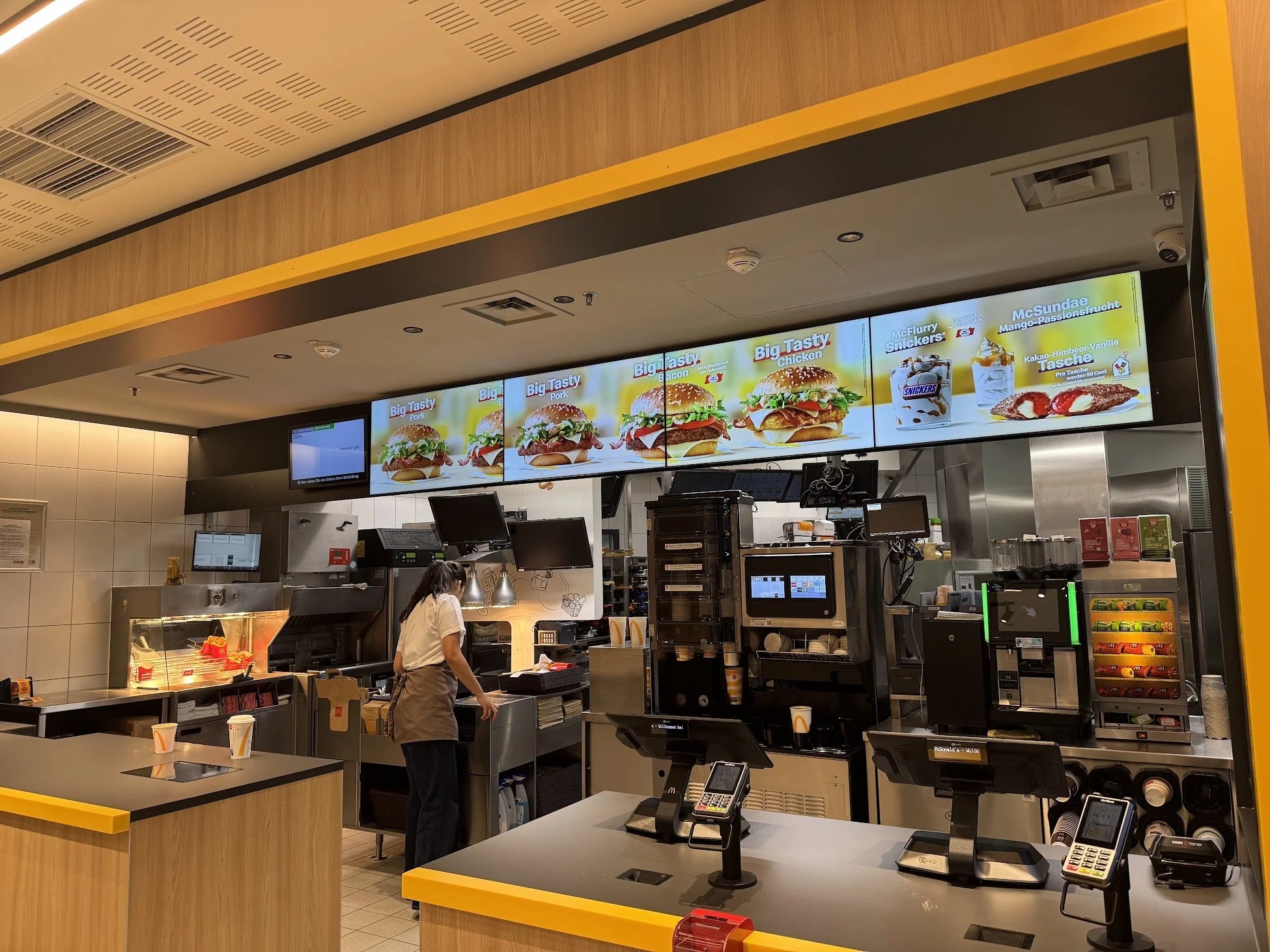 Inside of a fast-food restaurant with digital menu screens overhead displaying burger and dessert options, a cashier station with POS systems, and a worker behind the counter preparing orders.
