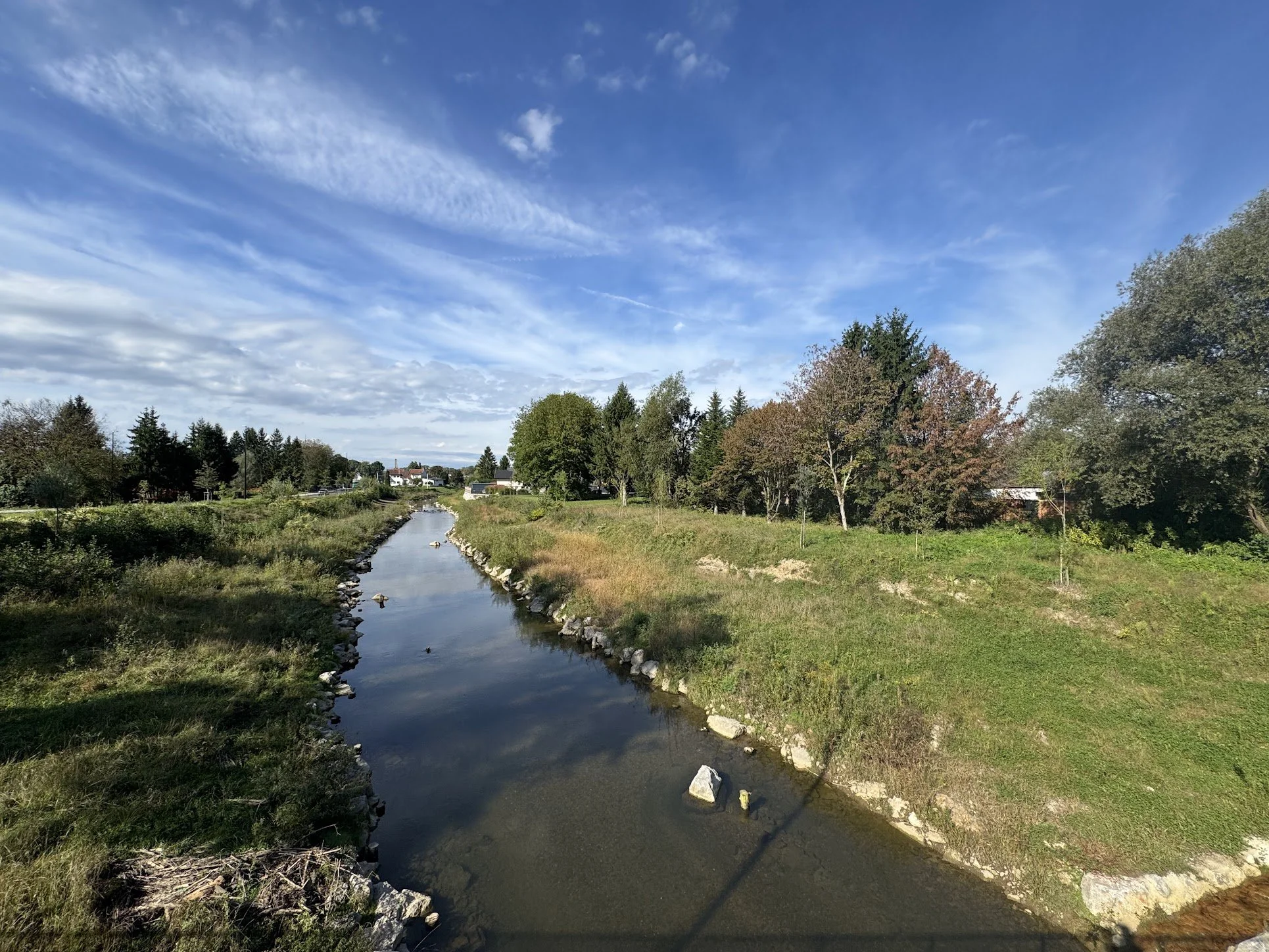 A small river or creek flowing through a grassy area with trees on either side, under a blue sky with wispy clouds.