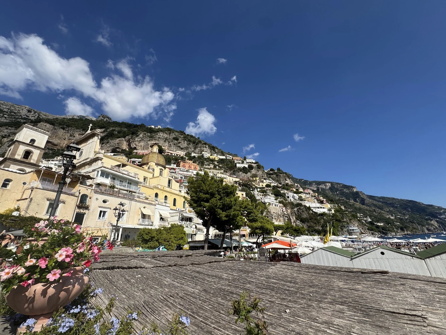 Colorful hillside buildings with a church and a blue sky with white clouds, overlooking a waterfront with boats and outdoor cafes.