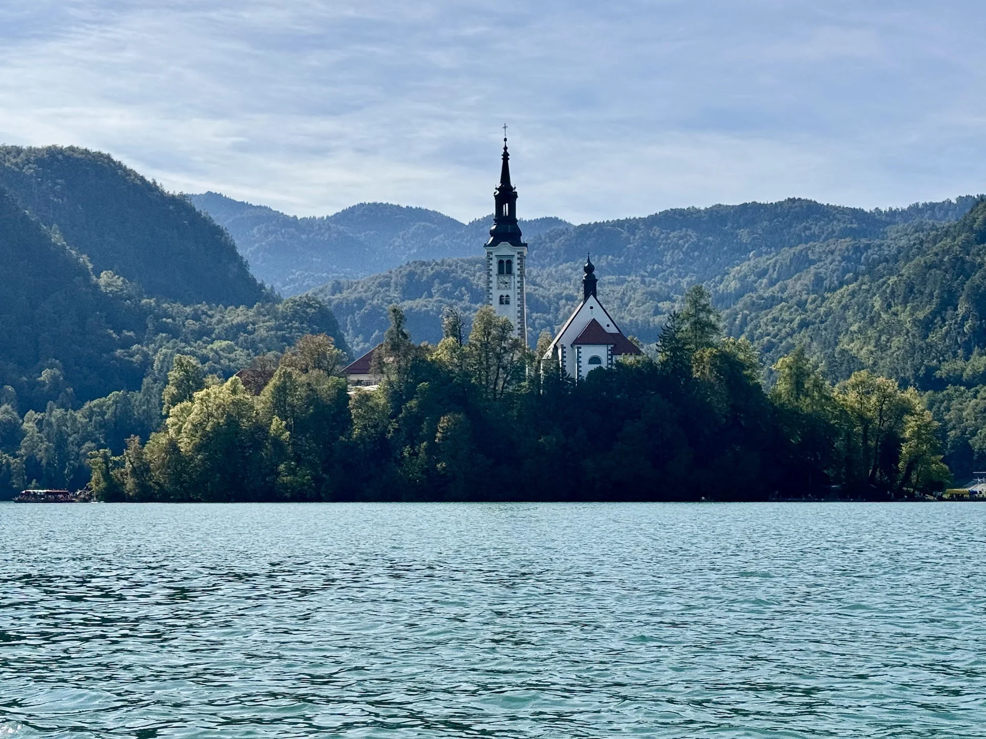 A church with a tall steeple located among trees and mountains, with a body of water in the foreground.