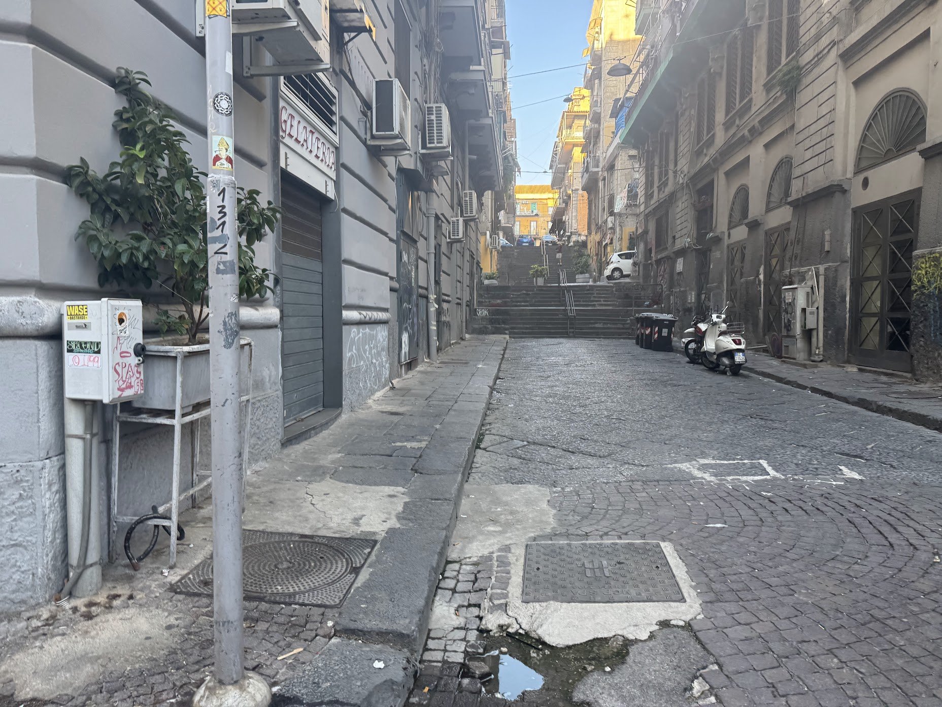 A narrow city street with cobblestone pavement and a flight of steps in the background. There are parked scooters and two trash bins on the side. Buildings on both sides have balconies and air conditioning units. A small puddle of water is visible in