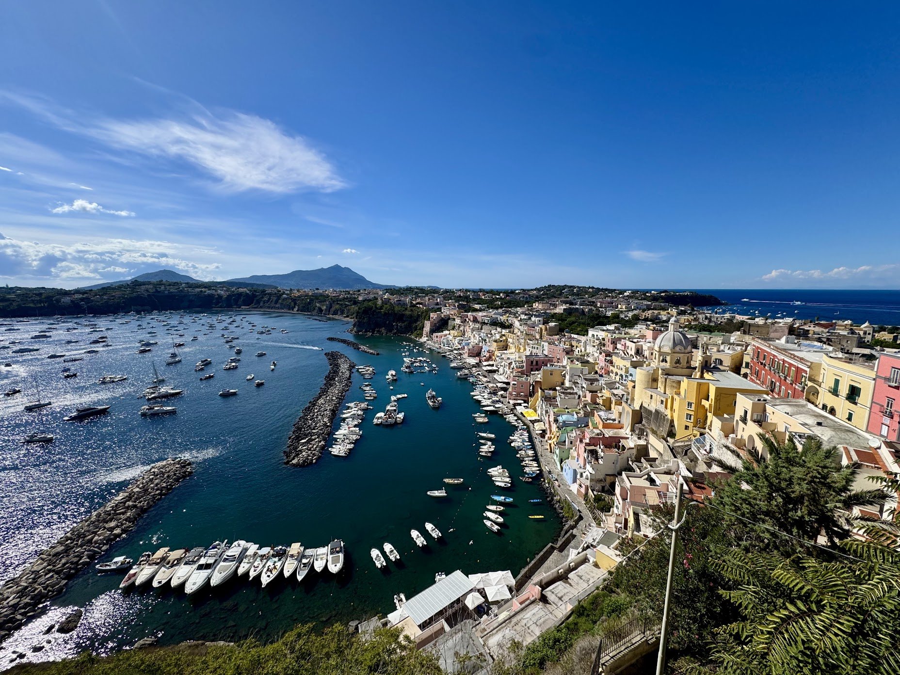 Scenic view of a coastal Italian town with colorful buildings, a harbor filled with boats, and mountainous landscape in the background on a clear, sunny day.