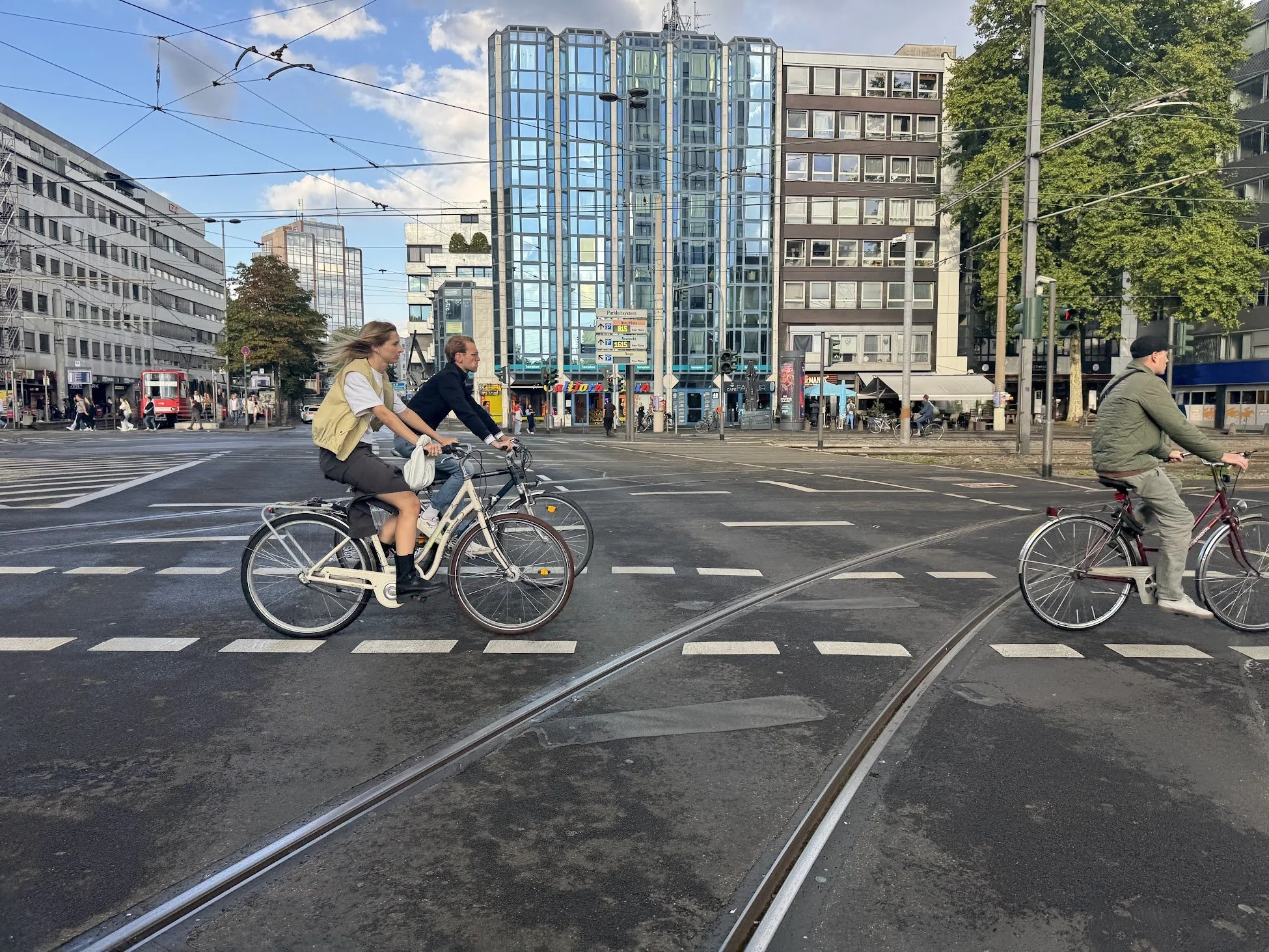 People riding bicycles across a city intersection with tram tracks, surrounded by tall buildings and trees, under a partly cloudy sky.