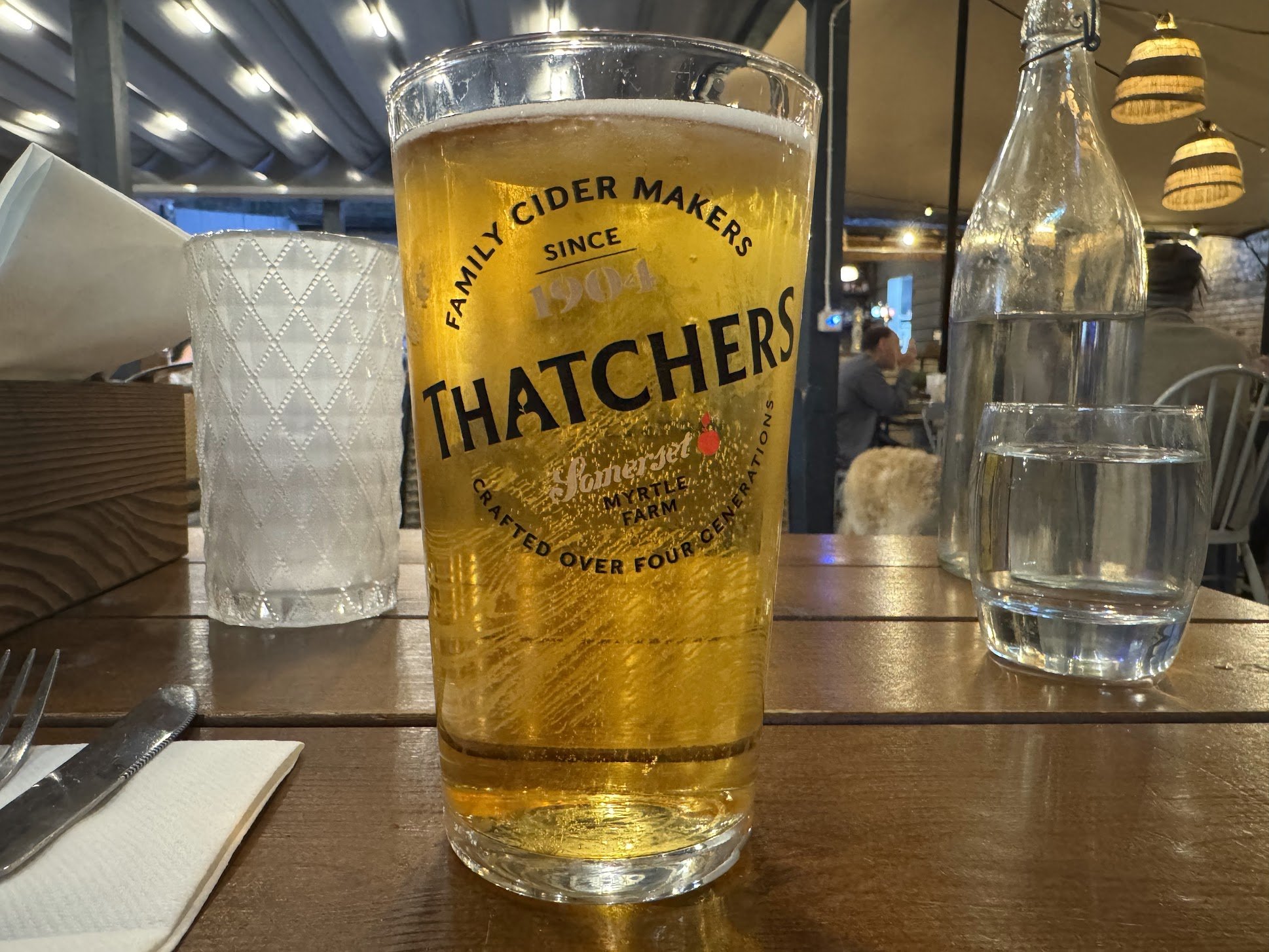 A tall glass of golden beer with the logo 'Thatchers' on it, placed on a wooden table in a restaurant setting. In the background, there are water bottles, a glass, a napkin holder, and some hanging lights.
