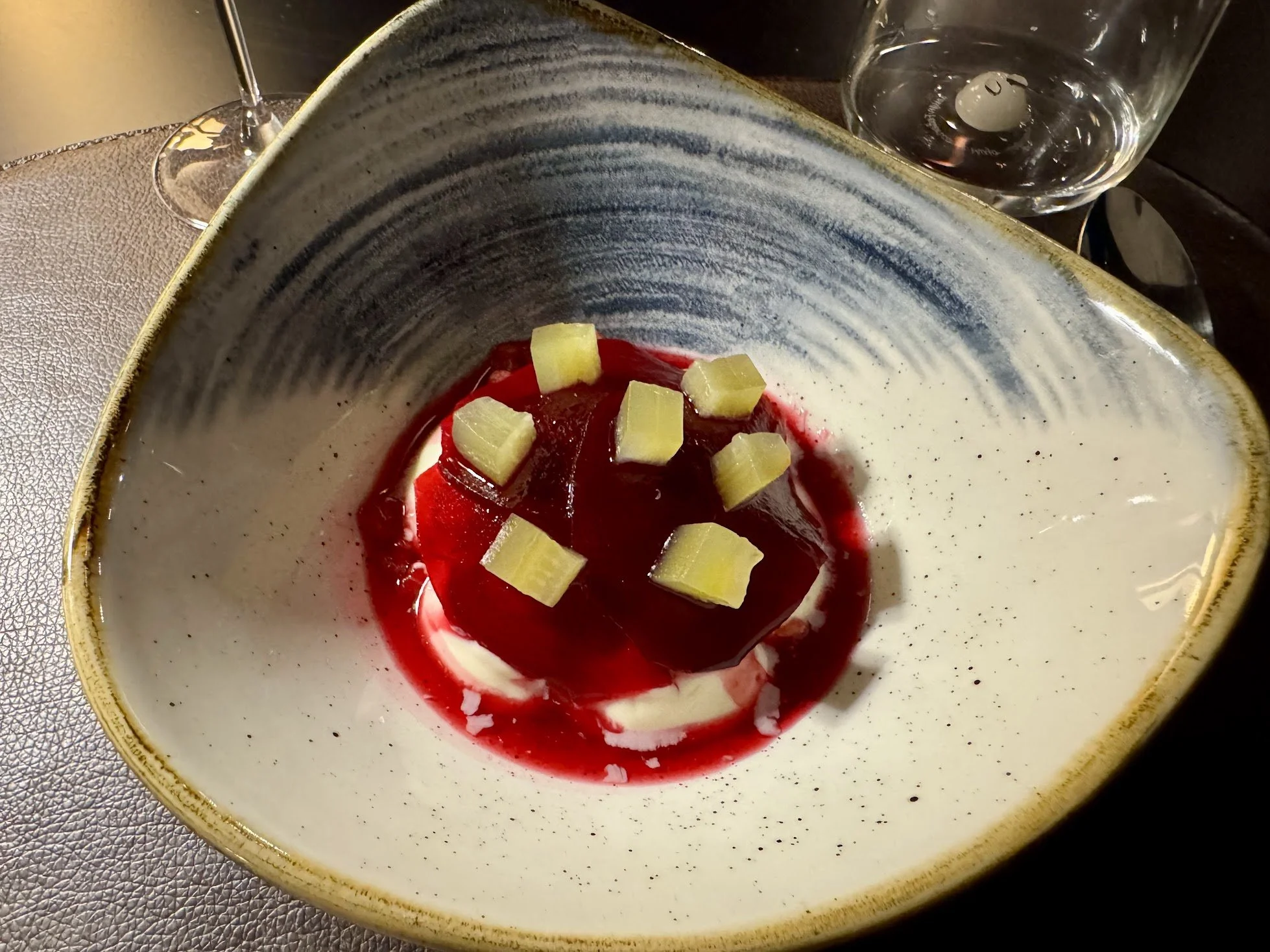 A ceramic bowl with a beige and dark blue design, containing a dessert with red jelly, white cream, and small yellow chunks, likely fruit or candy. A glass of water and a wine glass are visible on the table.