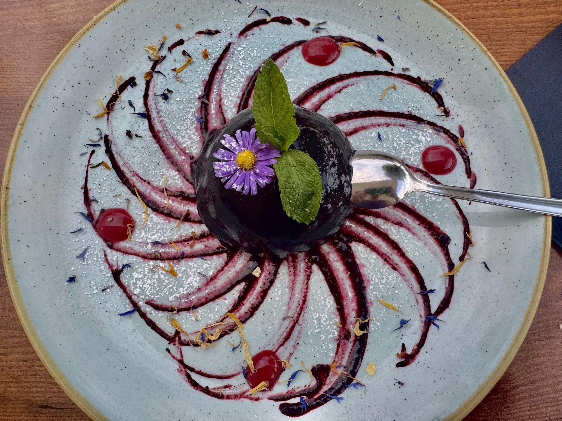 An elegant dessert plate with a scoop of dark purple sorbet garnished with a mint leaf and a purple flower, surrounded by artistic swirls of berry sauce and dotted with edible flower petals.