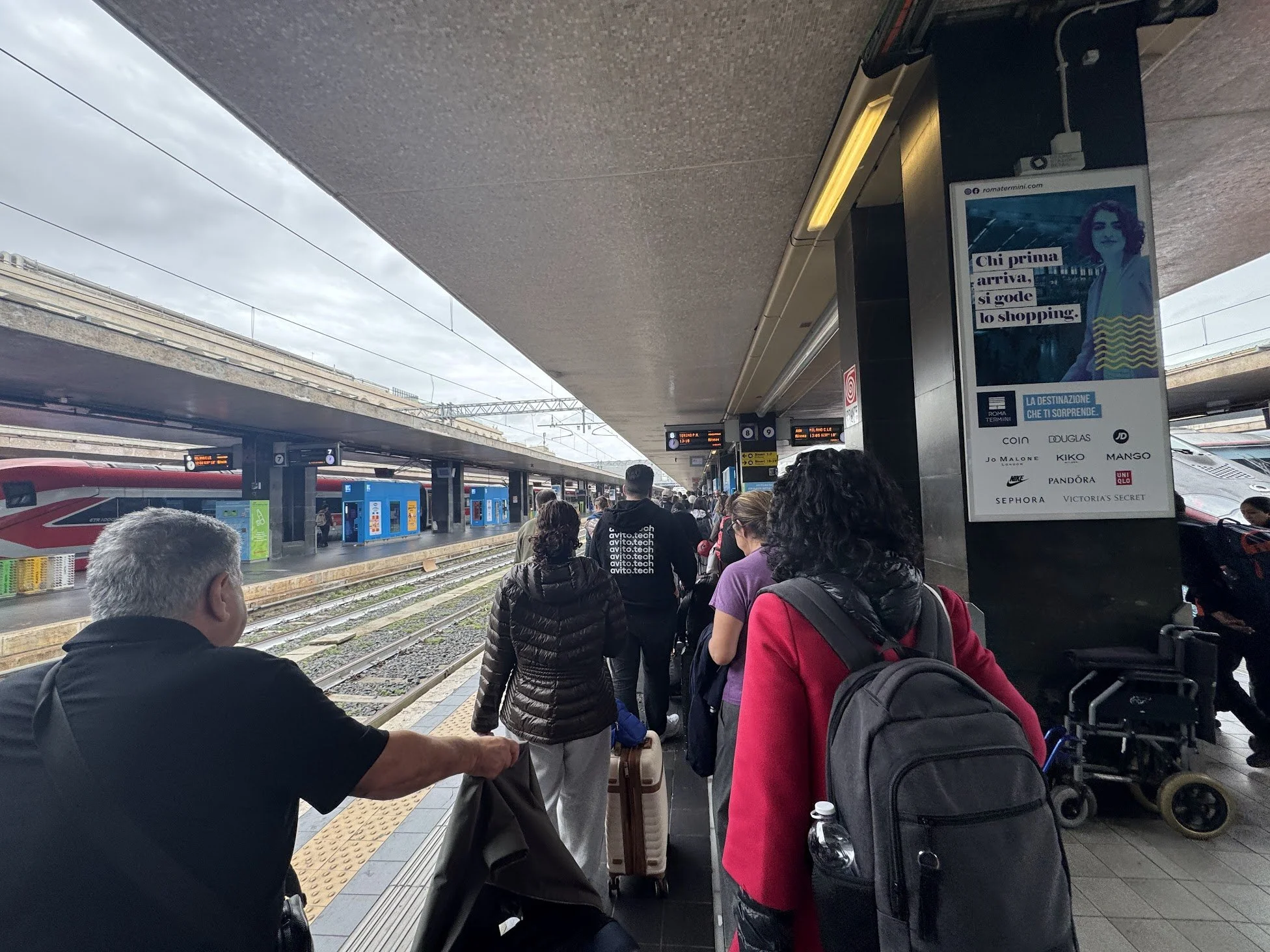 People waiting at a train station platform, some with luggage, under an overhang with trains on tracks and digital signs overhead.