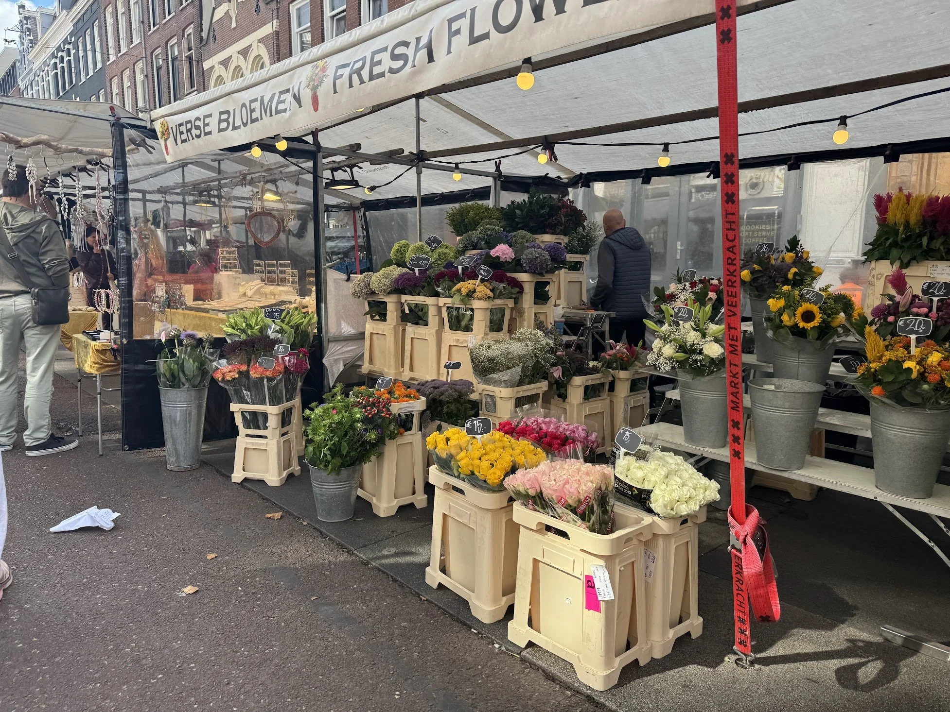 A flower stall on a city street with various colorful flowers including roses, tulips, and sunflowers, displayed in crates and buckets. The stand is shaded by a white canopy with a sign reading 'VERSE BLOEMEN FRESH FLO...' and lit with small yellow b