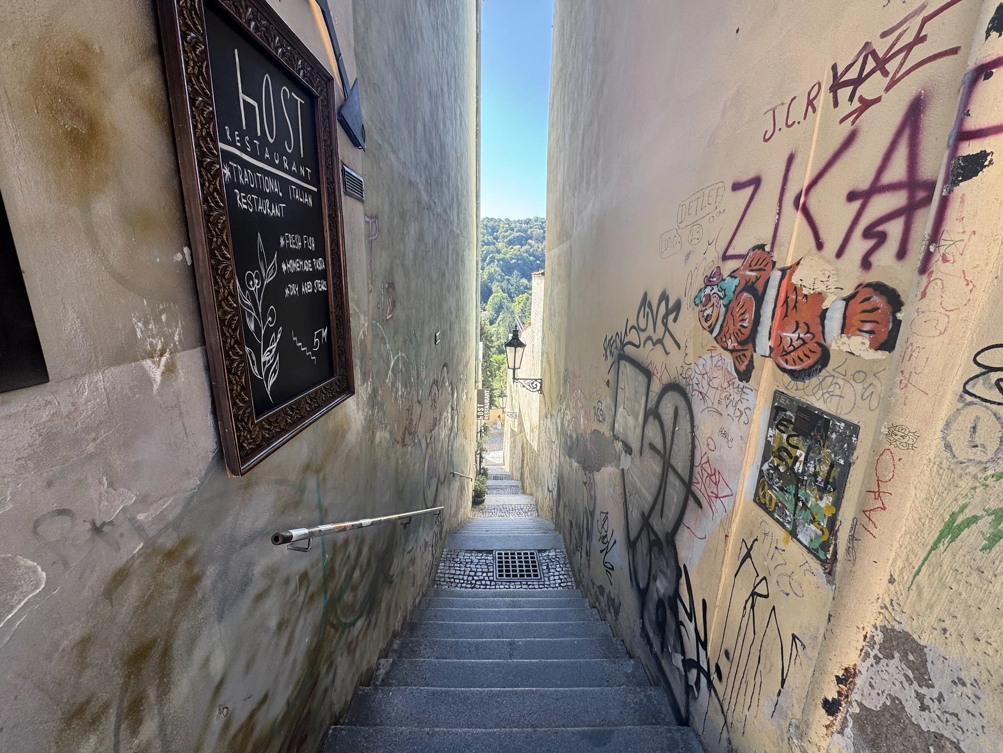 A narrow outdoor passageway between two walls. The left wall has some graffiti and a decorative framed blackboard sign that reads 'HOST RESTAURANT TRADITIONAL ITALIAN RESTAURANT' with menu items below. The right wall has graffiti and a cartoonish drawing of a tiger. The ground is covered in colorful graffiti and stickers. In the distance, a small hill with trees and a building with a sloped roof is visible, and the sky is clear and blue.