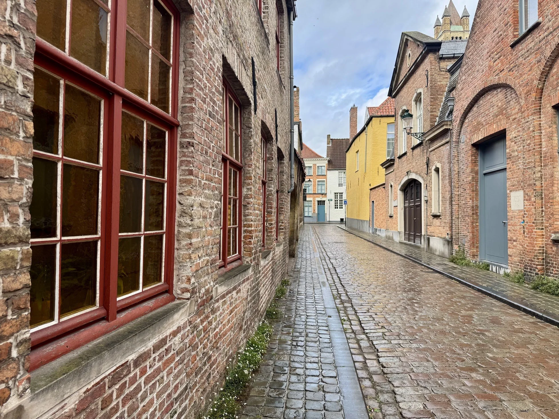 A narrow cobblestone street lined with old brick buildings, some with colorful painted facades, and decorative street lamps, under a cloudy sky.