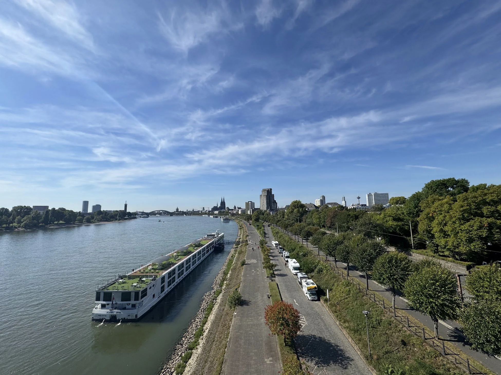 A cityscape with a river, tall buildings, and a boat docked along the quay. Trees line the waterfront and a clear blue sky overhead.