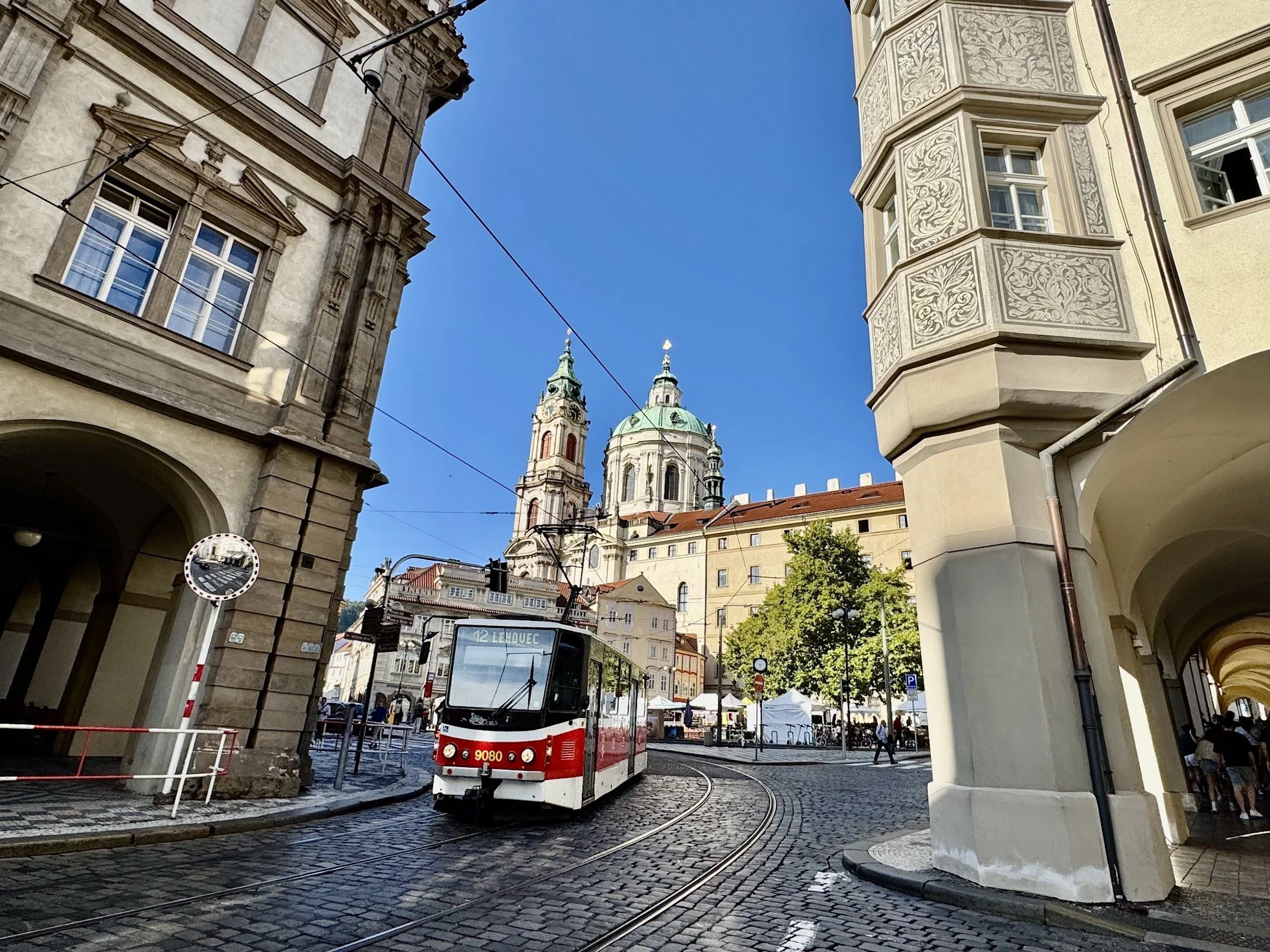 City street scene featuring a white and red tram on cobblestone tracks, historic buildings with detailed facades, and a church with green domes and clock towers in the background under a clear blue sky.