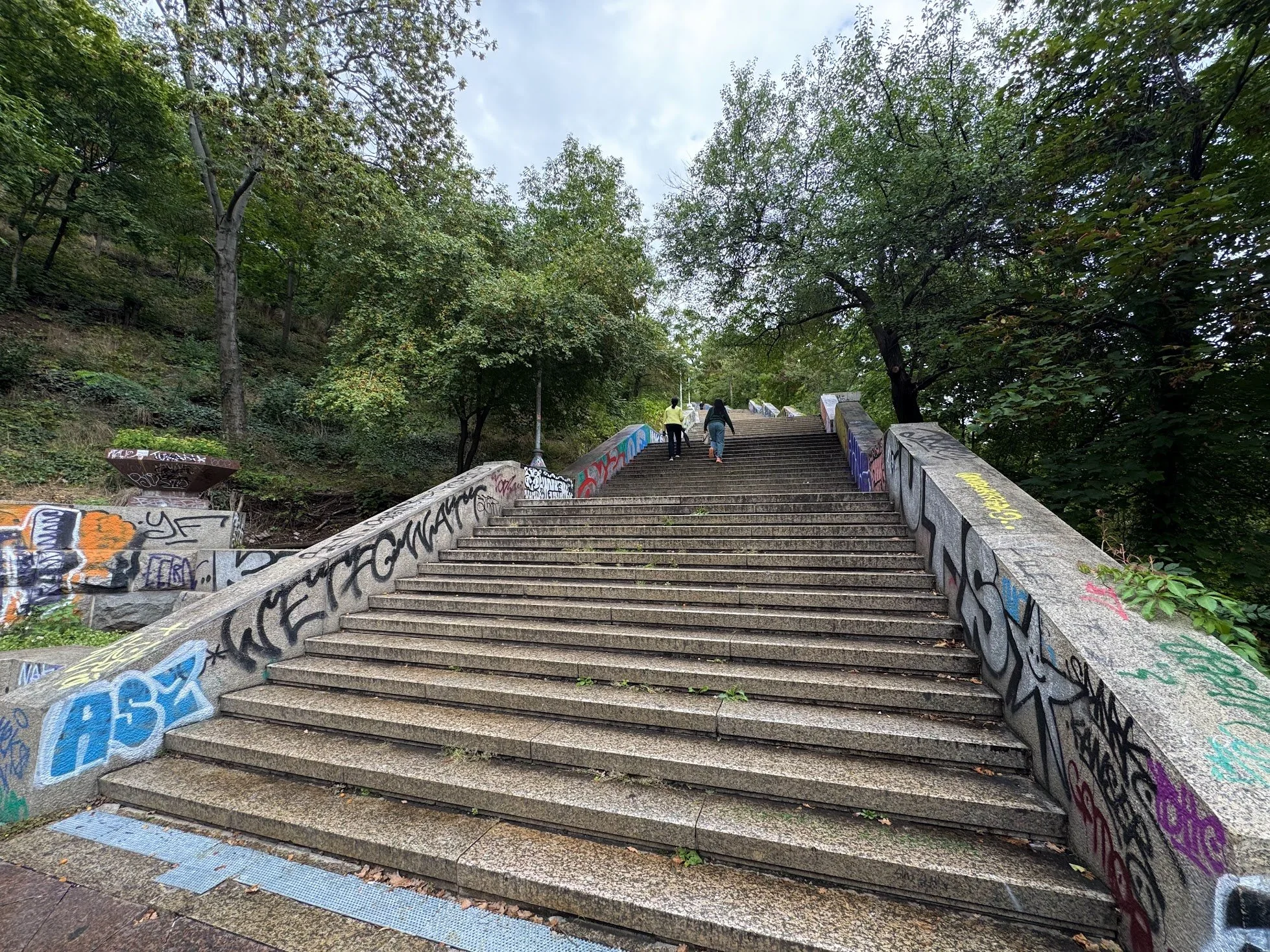 Concrete outdoor staircase covered in colorful graffiti, surrounded by lush green trees, with three people ascending the stairs on an overcast day.
