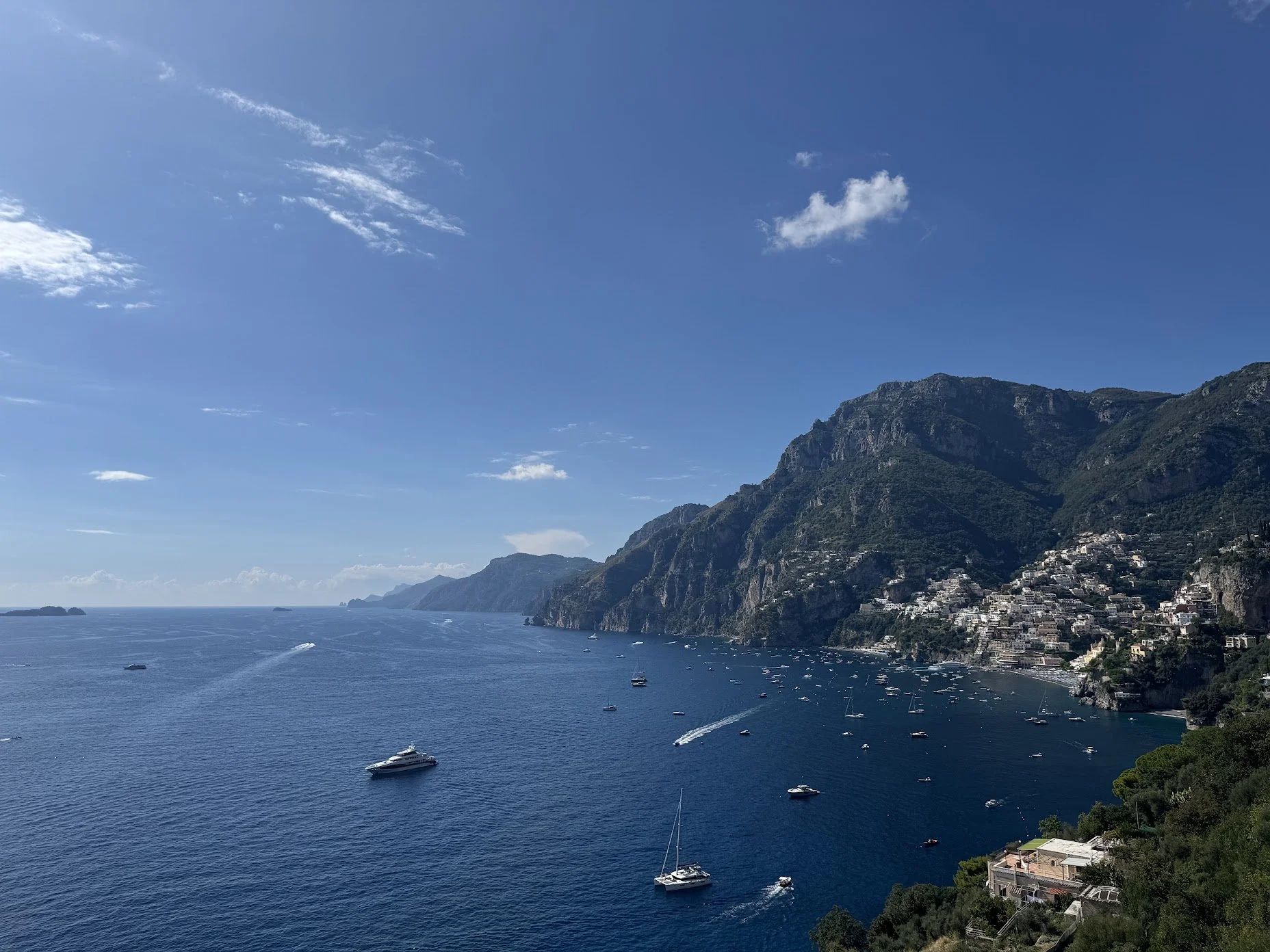 Scenic view of a coastal town with houses on the hillside, a clear blue sea with boats, and mountain cliffs under a partly cloudy sky.