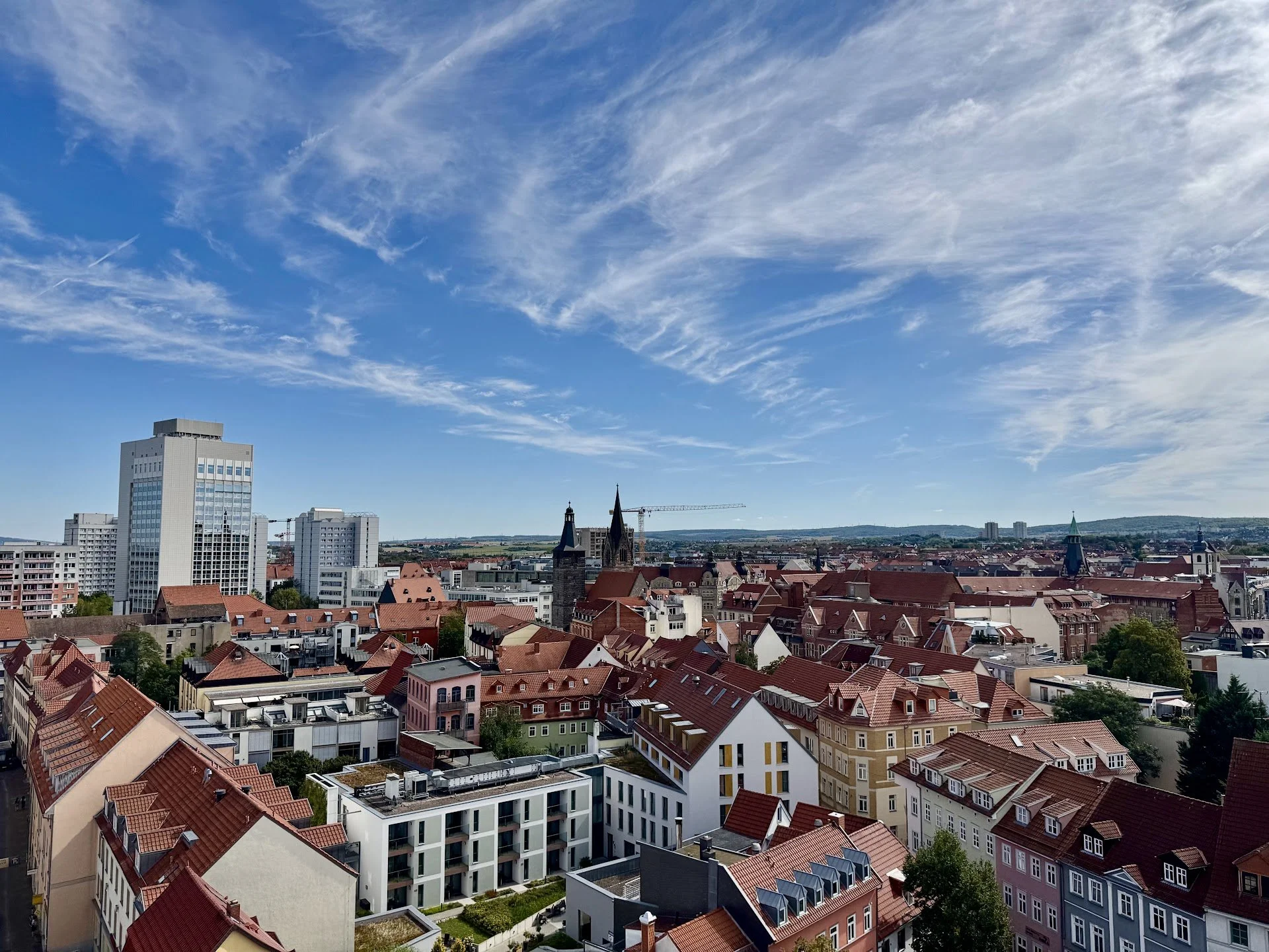 Cityscape with modern and historic buildings, red rooftops, tall skyscrapers, and a blue sky with wispy clouds.