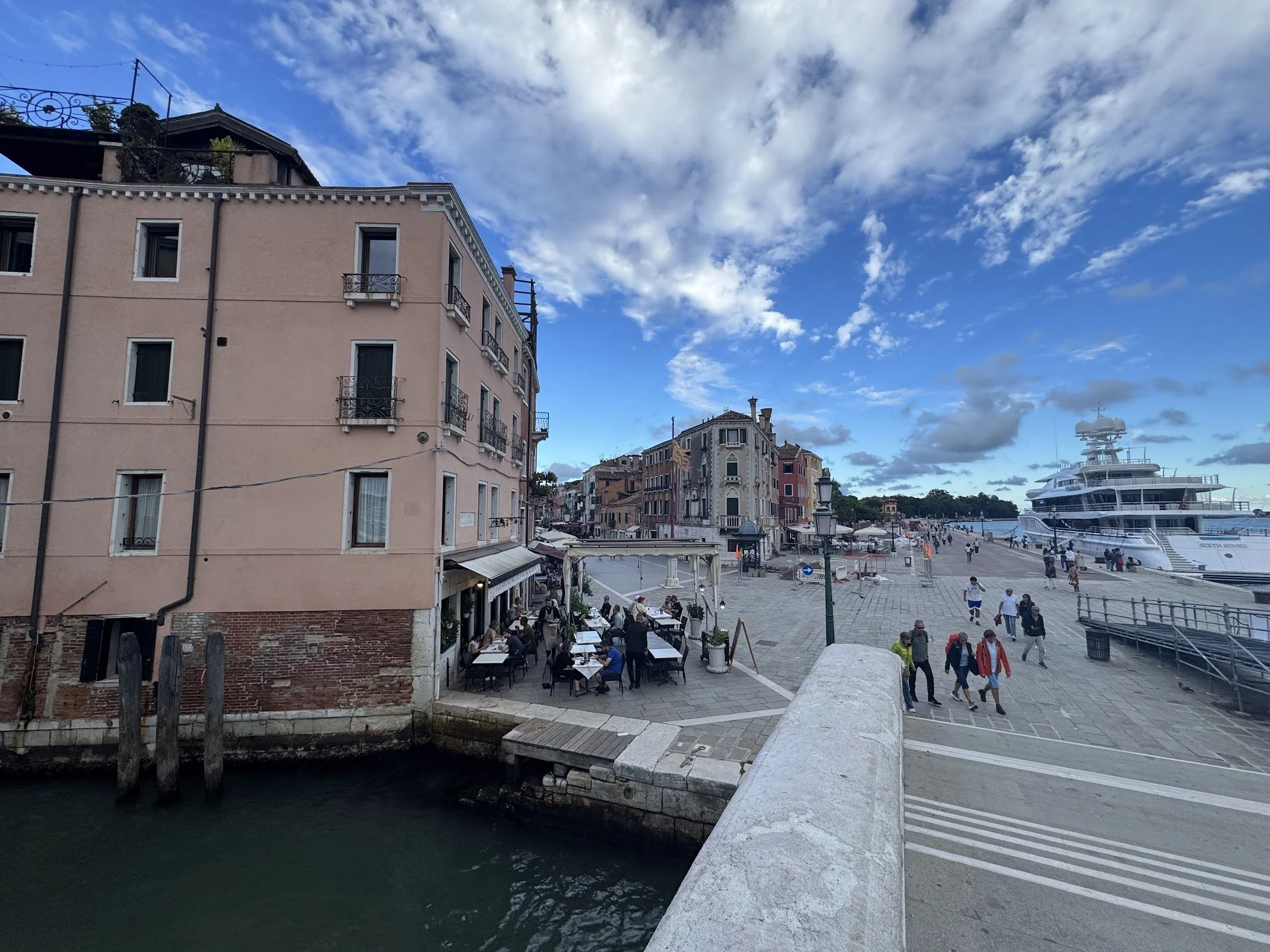 A coastal city scene featuring pastel-colored buildings, a crowded waterfront promenade with people walking and dining at outdoor tables, and a large yacht docked by the pier. The sky is partly cloudy with patches of blue.