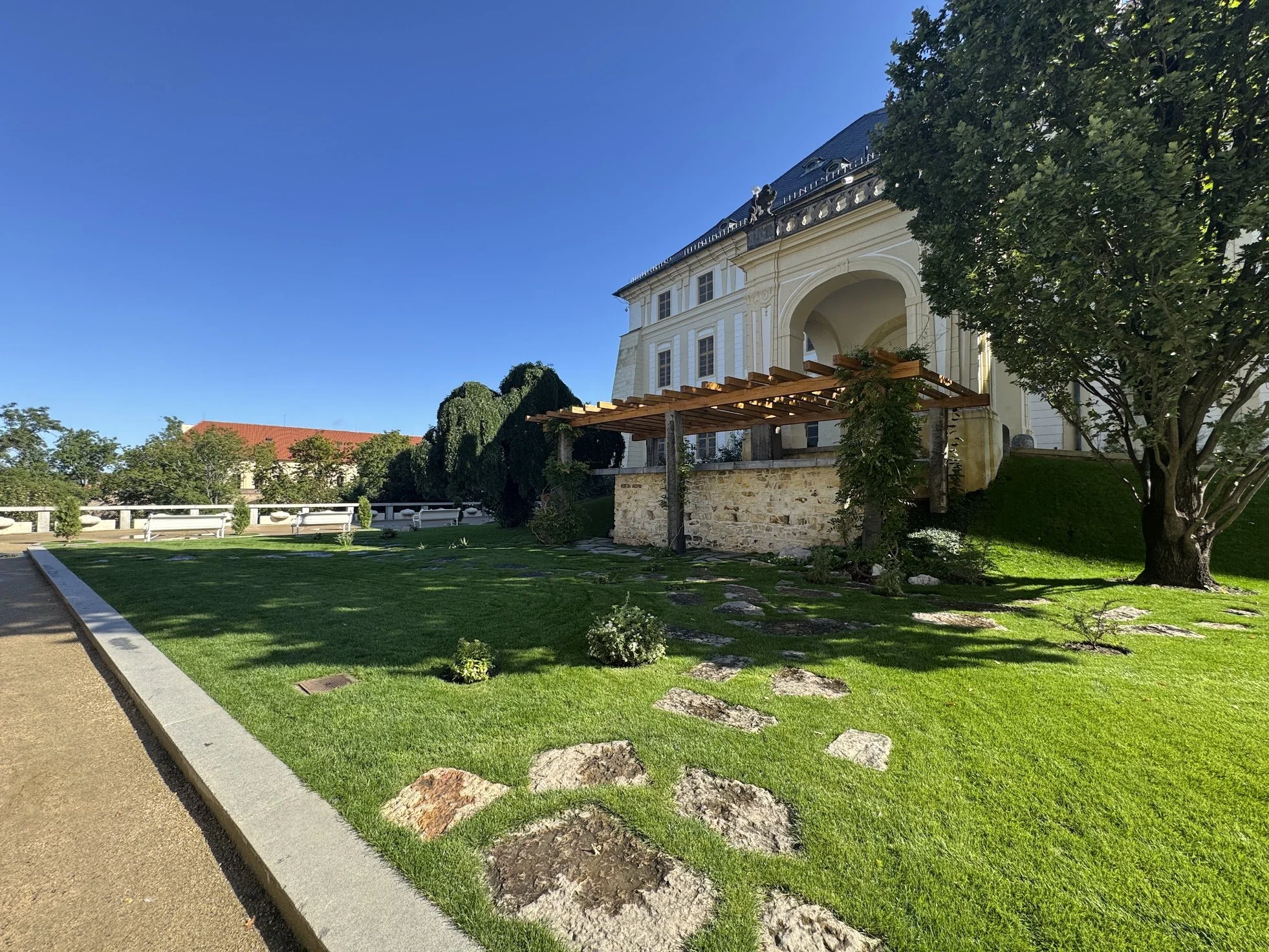 A historic white building with arched windows and a slate roof, surrounded by a landscaped garden with a gravel pathway, trimmed bushes, and a large tree, under a clear blue sky.